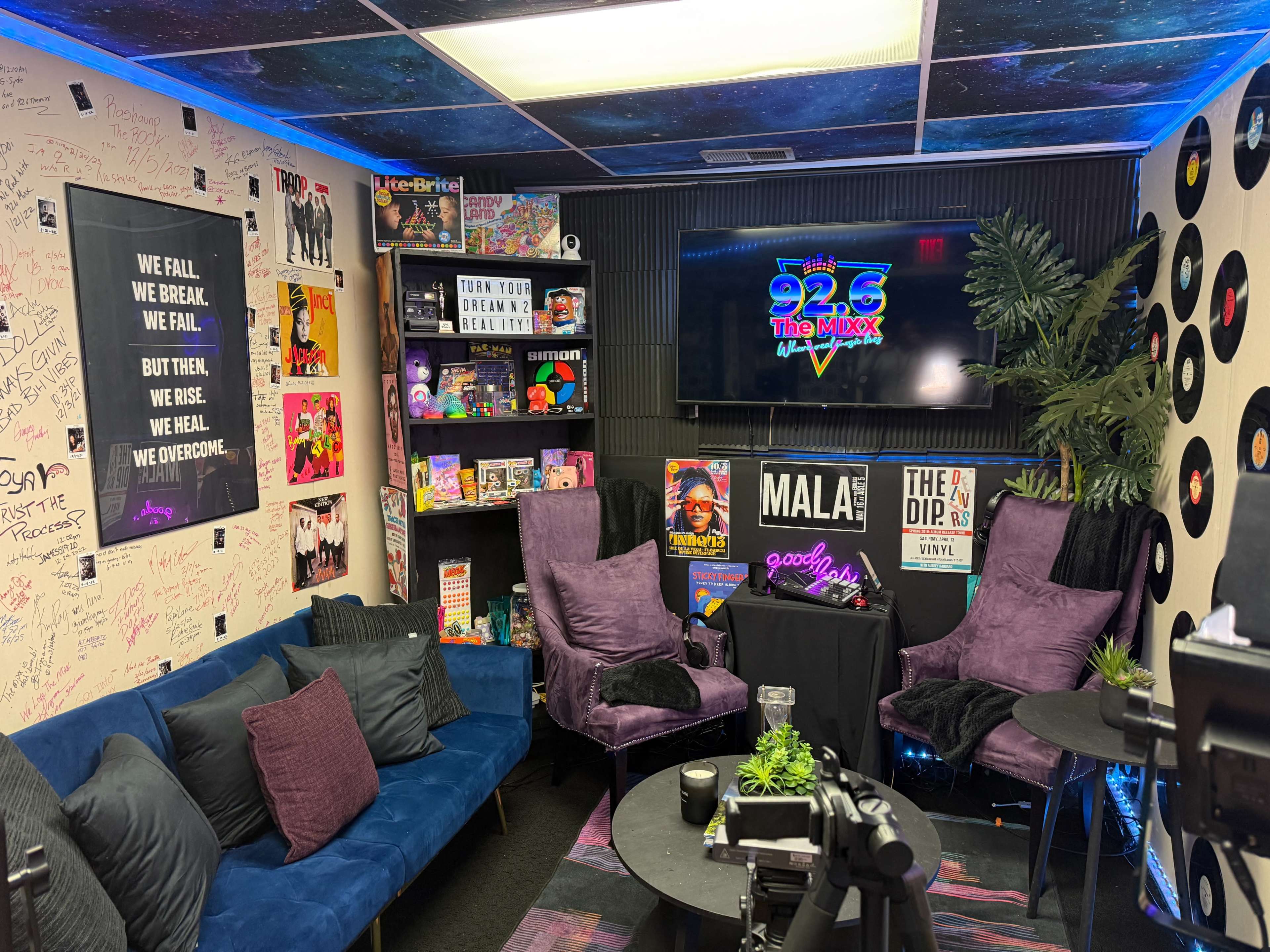 The image shows a vibrant, decorated studio space with a seating area featuring purple chairs and a blue couch, surrounded by colorful wall art and displays, along with a screen displaying "92.6 The Mixx."