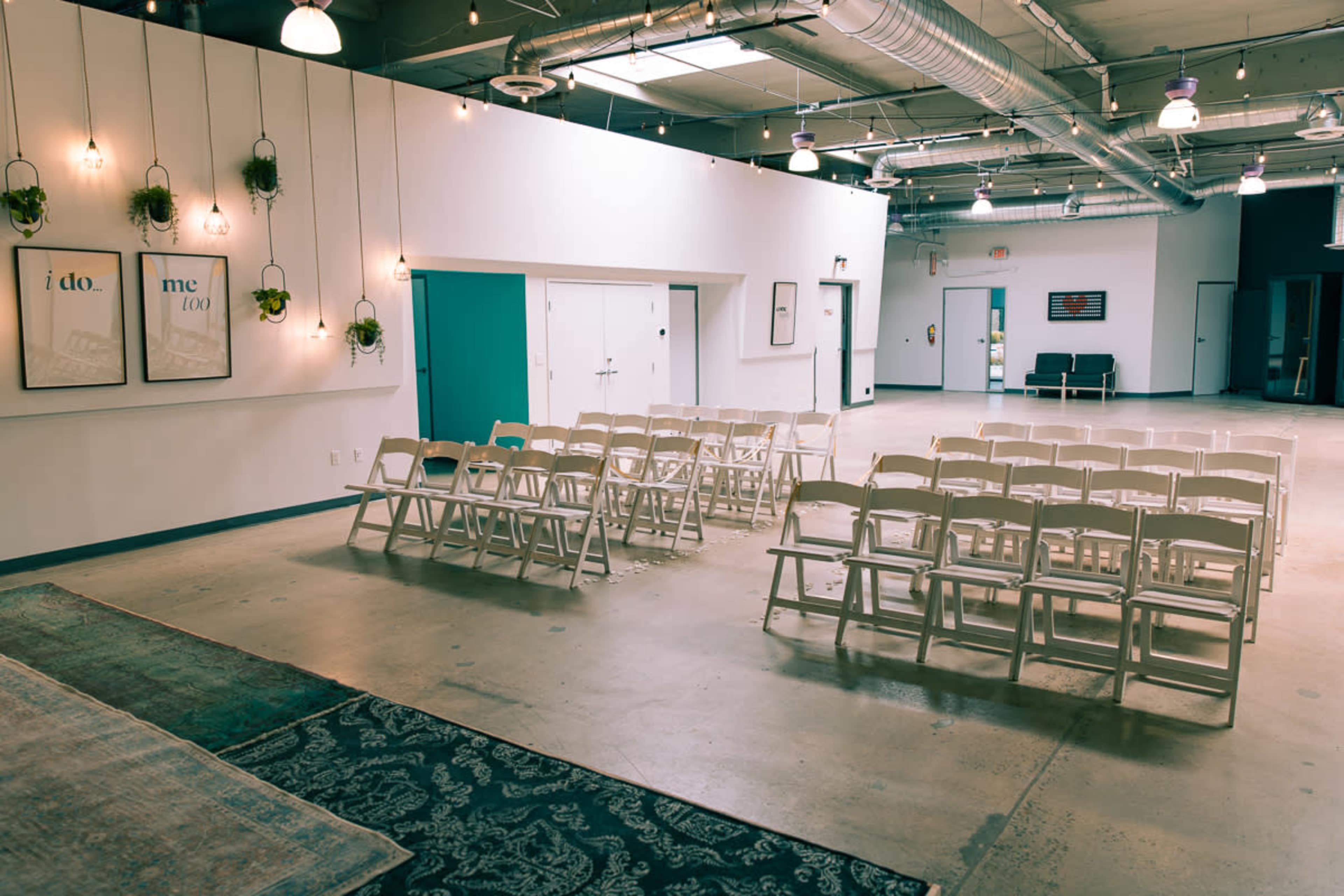 A spacious room set up for an event, featuring rows of white chairs arranged in front of a wall adorned with decorative plants and framed artwork.