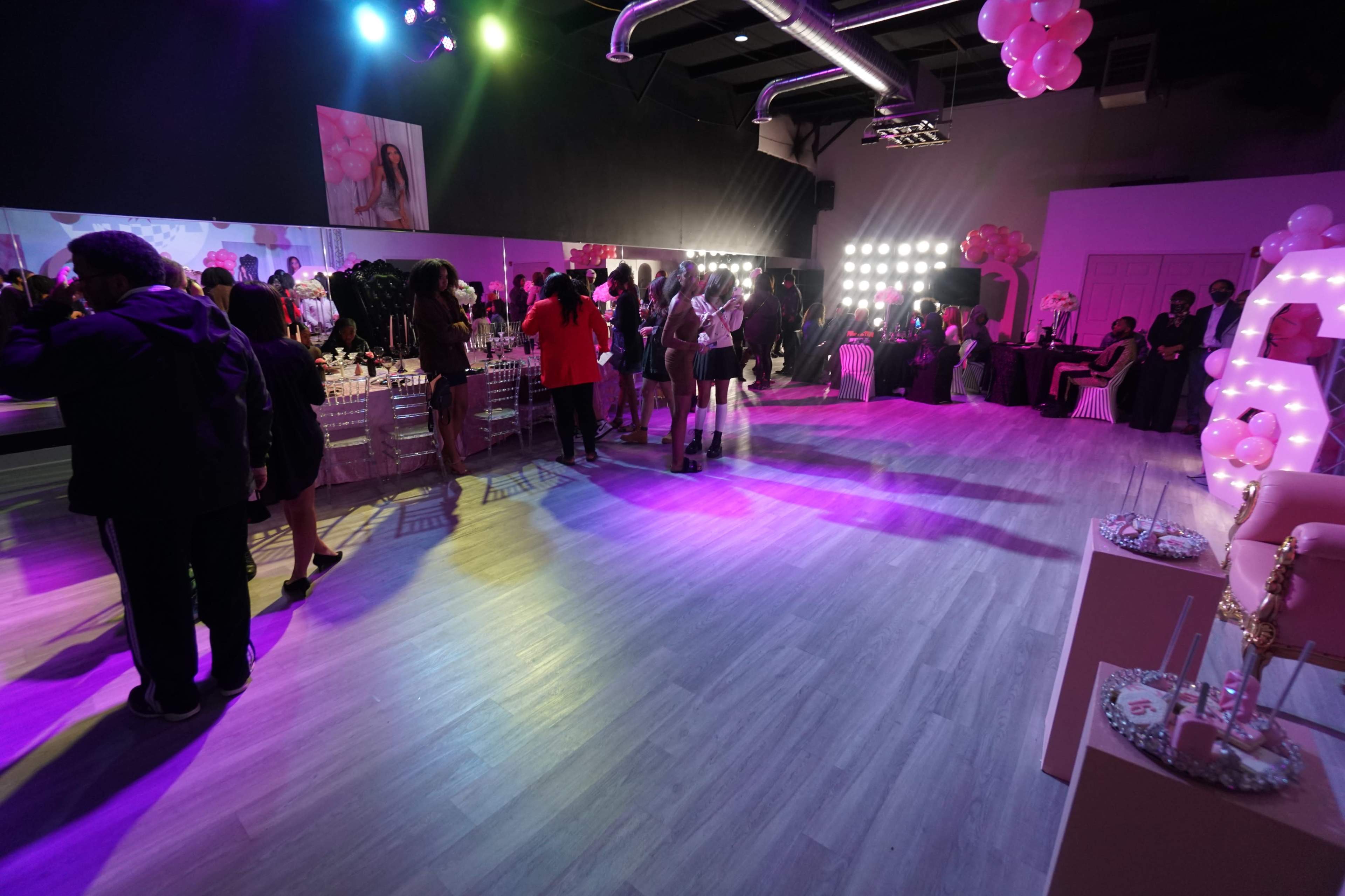 A crowded event space is filled with people socializing under colorful lighting, with tables set up for a celebration on one side.