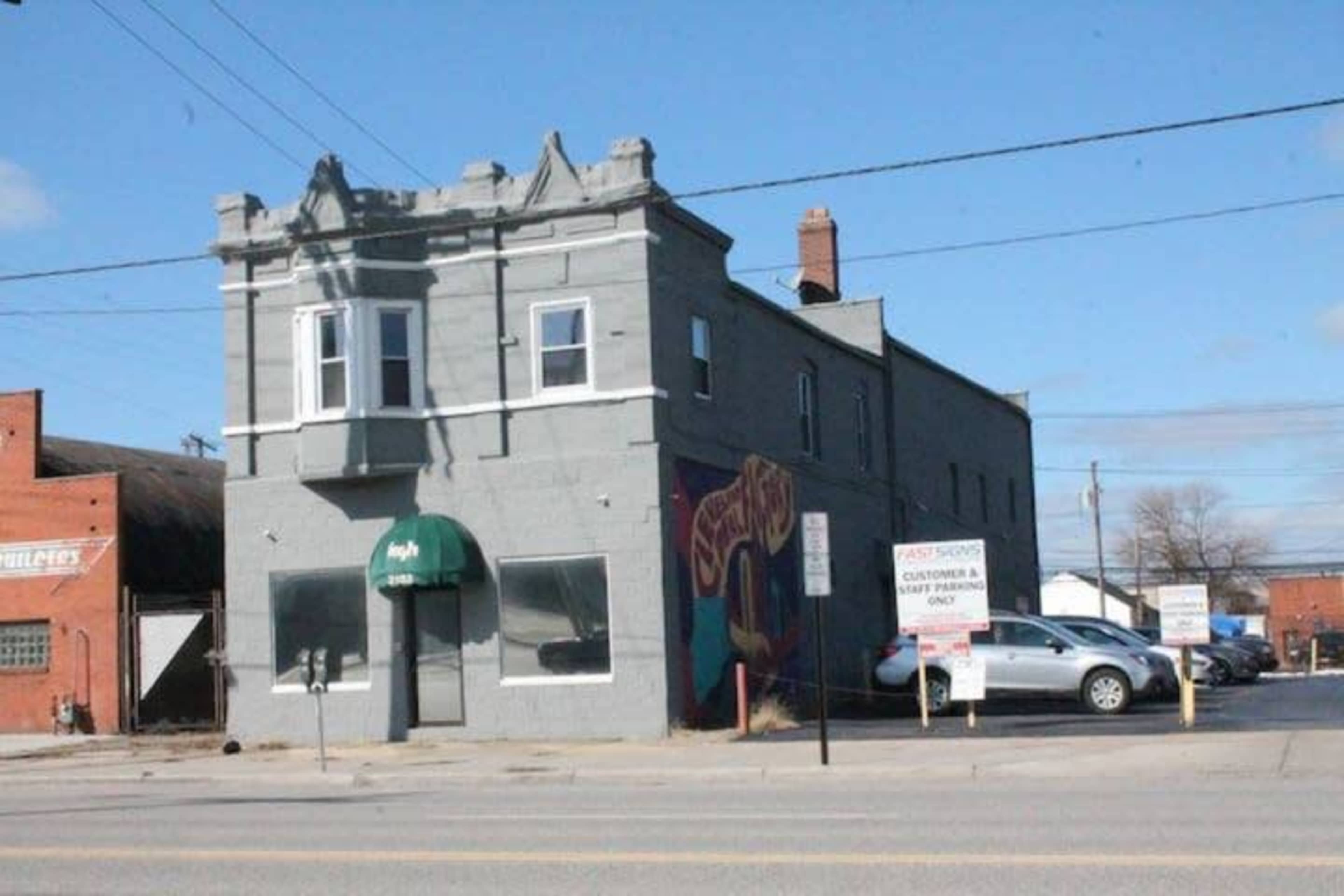 A two-story gray building with a green awning and a colorful mural on its side, positioned next to a parking lot and street signs.