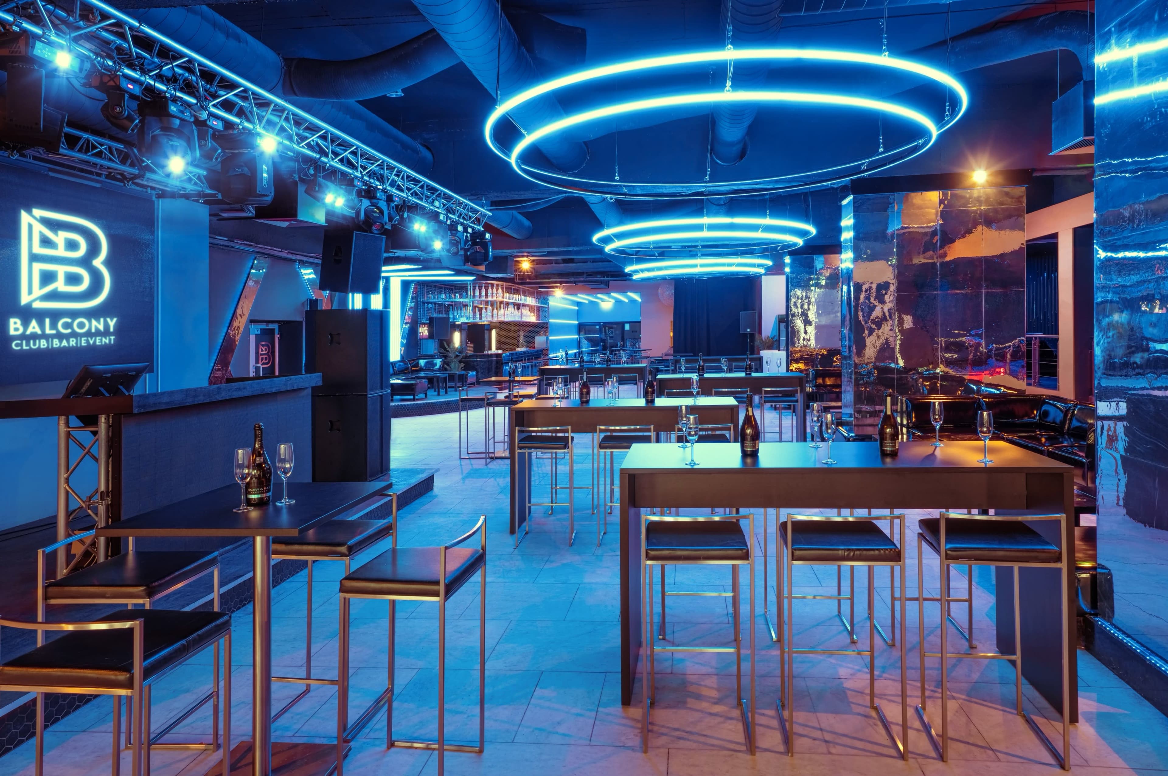 A modern nightclub interior featuring illuminated circular light fixtures, sleek tables, and a DJ booth with a large logo.
