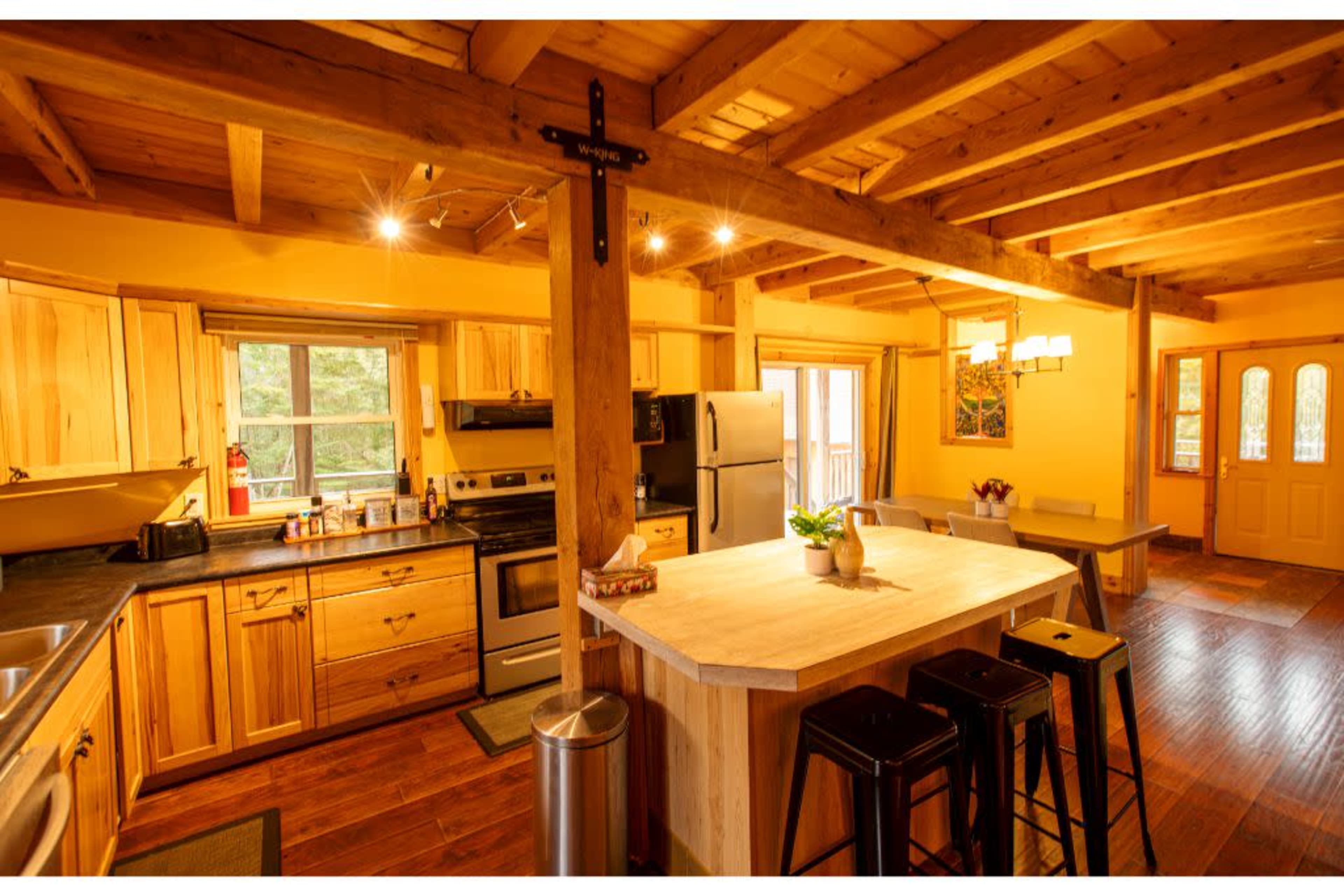 The image shows a wooden kitchen with yellow walls, featuring a central island, stainless steel appliances, and wooden cabinetry.