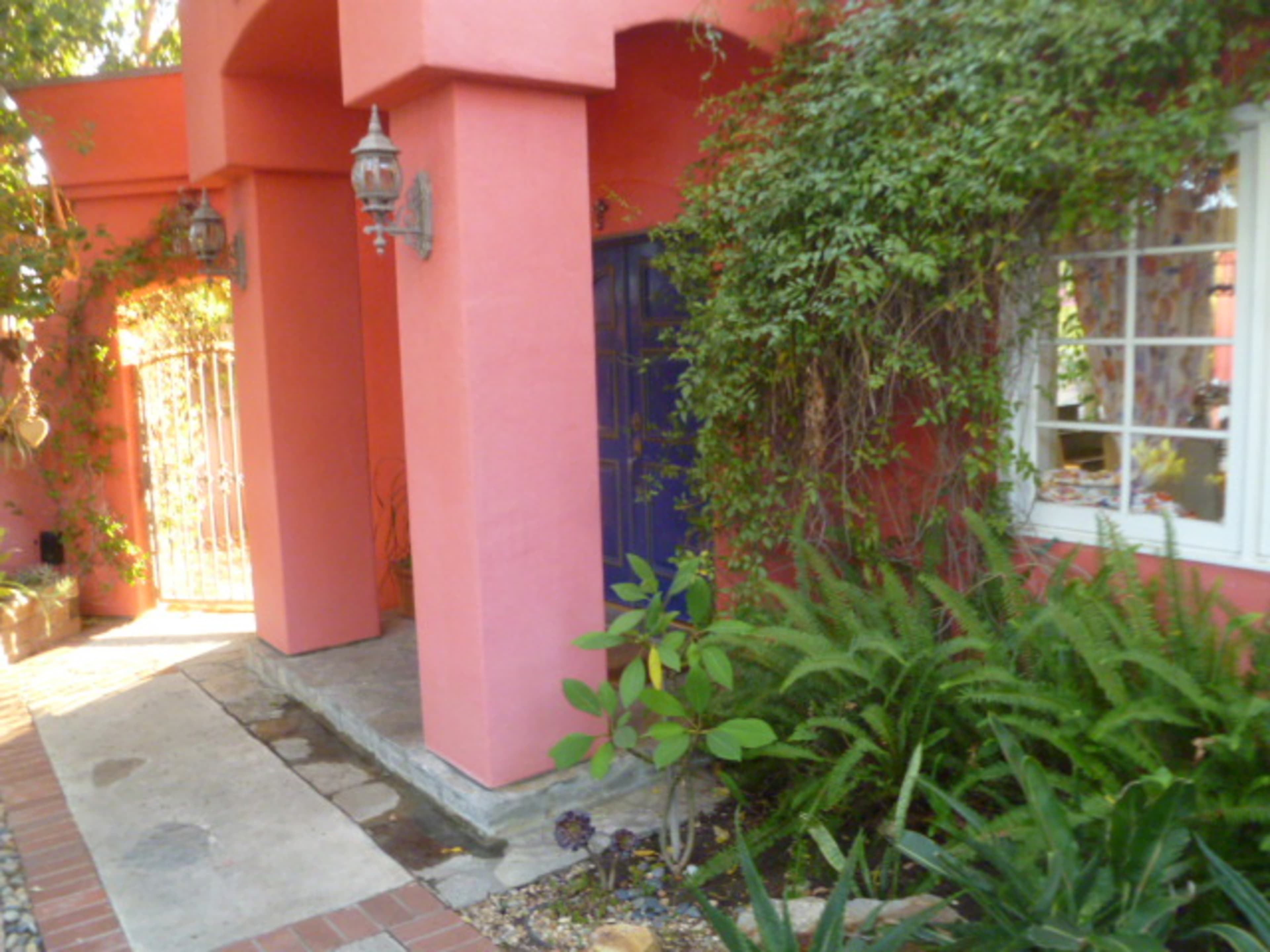 A vibrant pink house entrance with a blue door, bordered by lush greenery and a stone pathway.