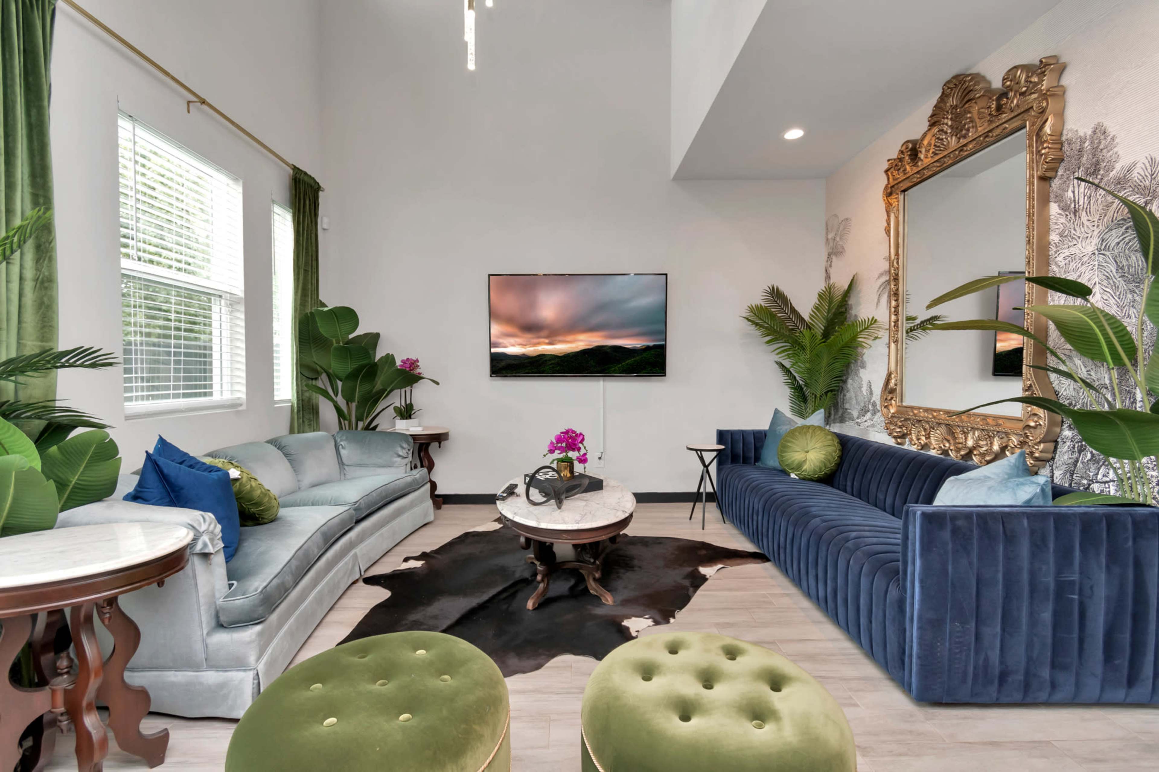 A stylish living room featuring a blue velvet sofa, a gray couch, a round coffee table, green plants, and a large golden mirror above the television.