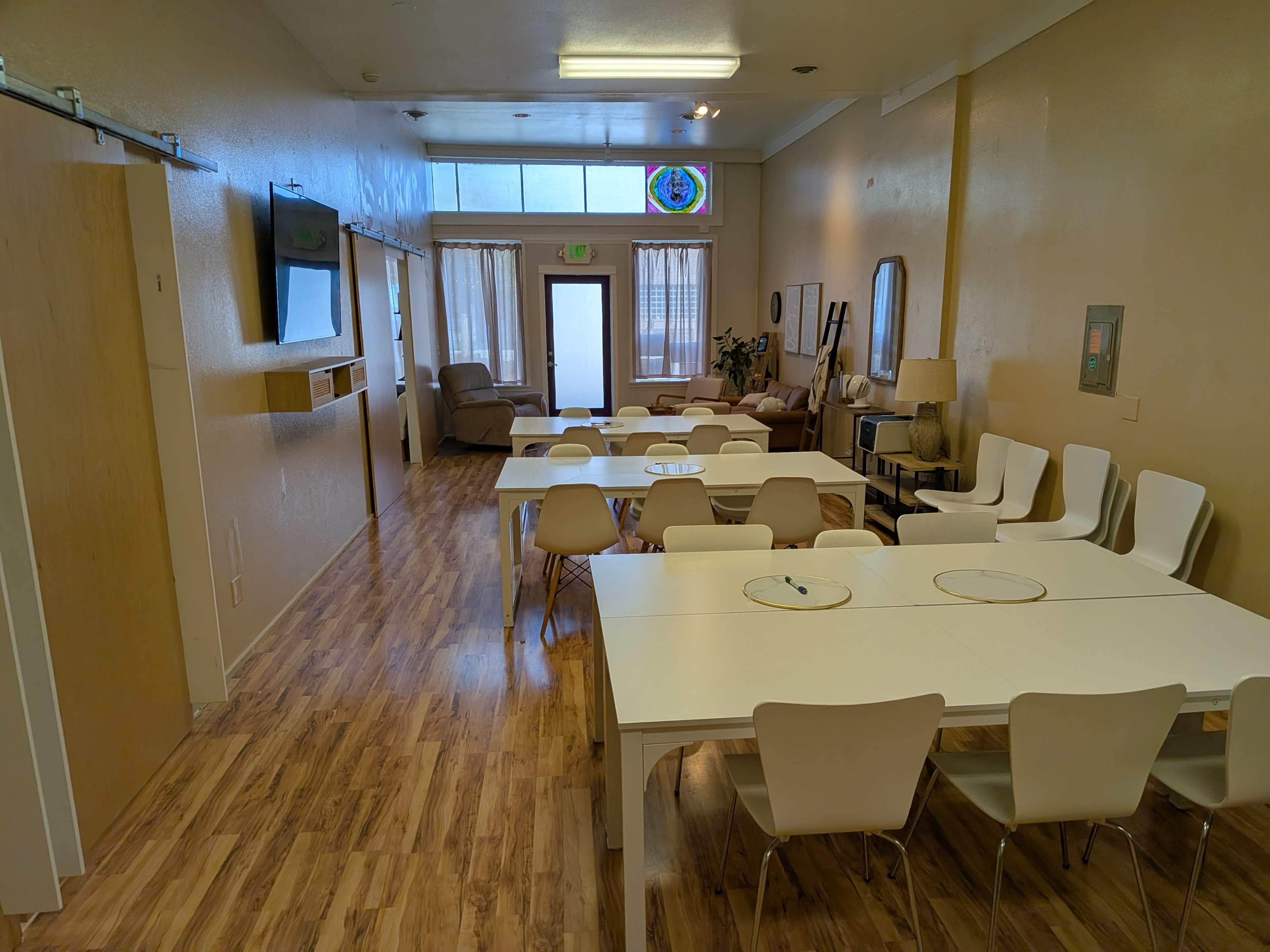 The image shows a spacious room with multiple white tables and chairs arranged neatly, two armchairs in the background, and large windows allowing light to enter.