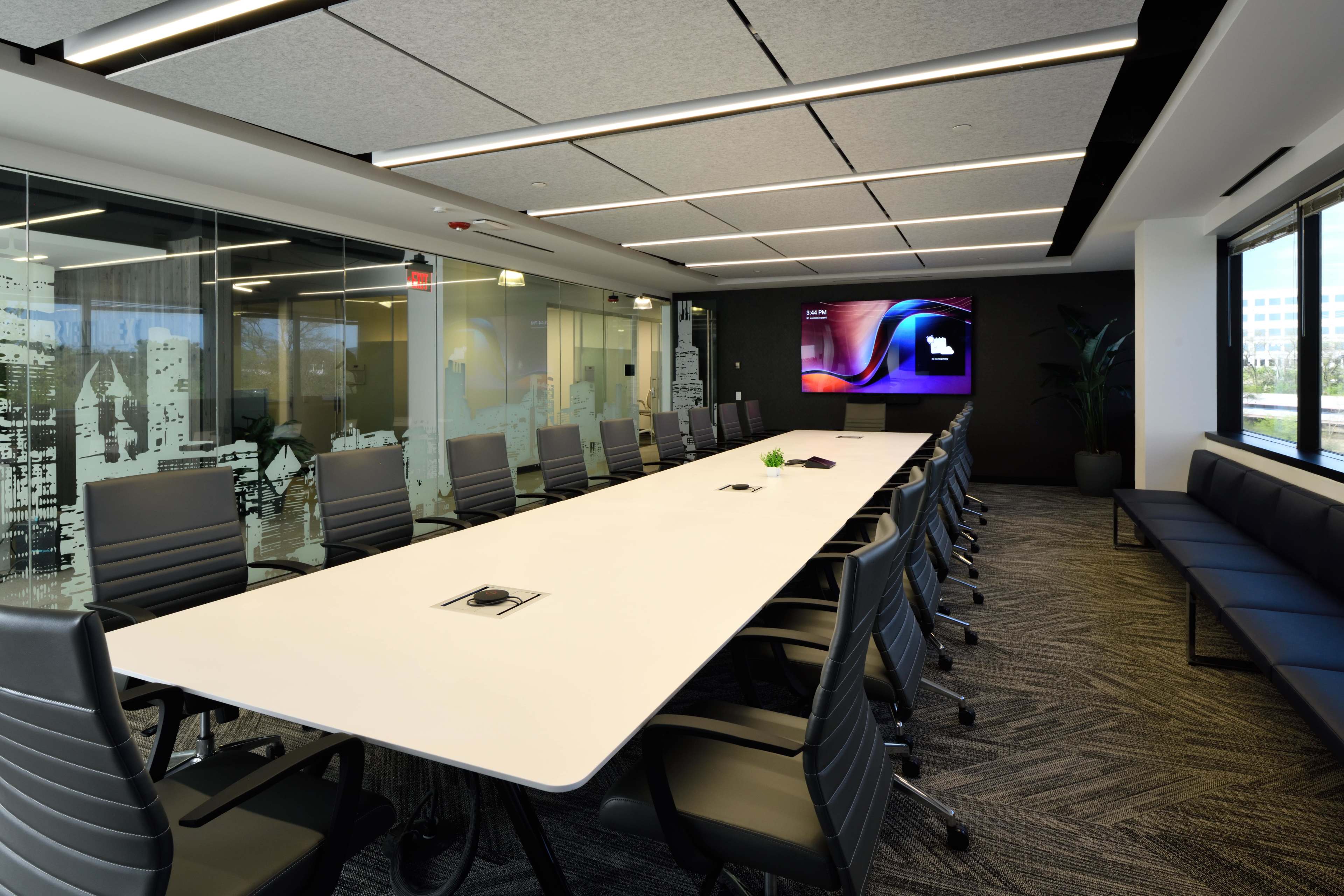 A modern conference room features a long white table surrounded by ergonomic chairs, with a large screen mounted on the wall and glass panels adorned with cityscape graphics.