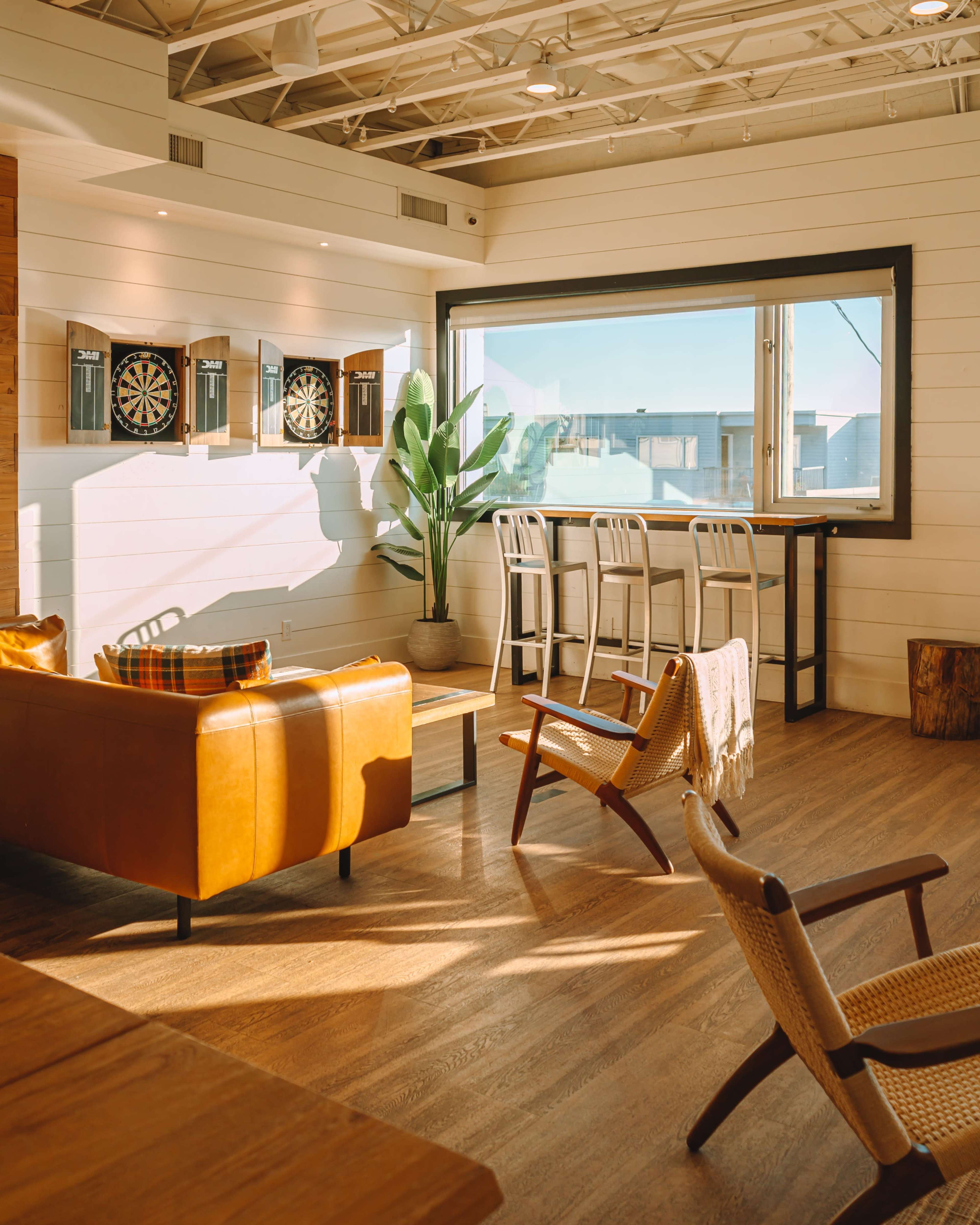 A modern, airy room with a leather sofa, a wooden table, bar stools by a window, and dartboards on the wall.