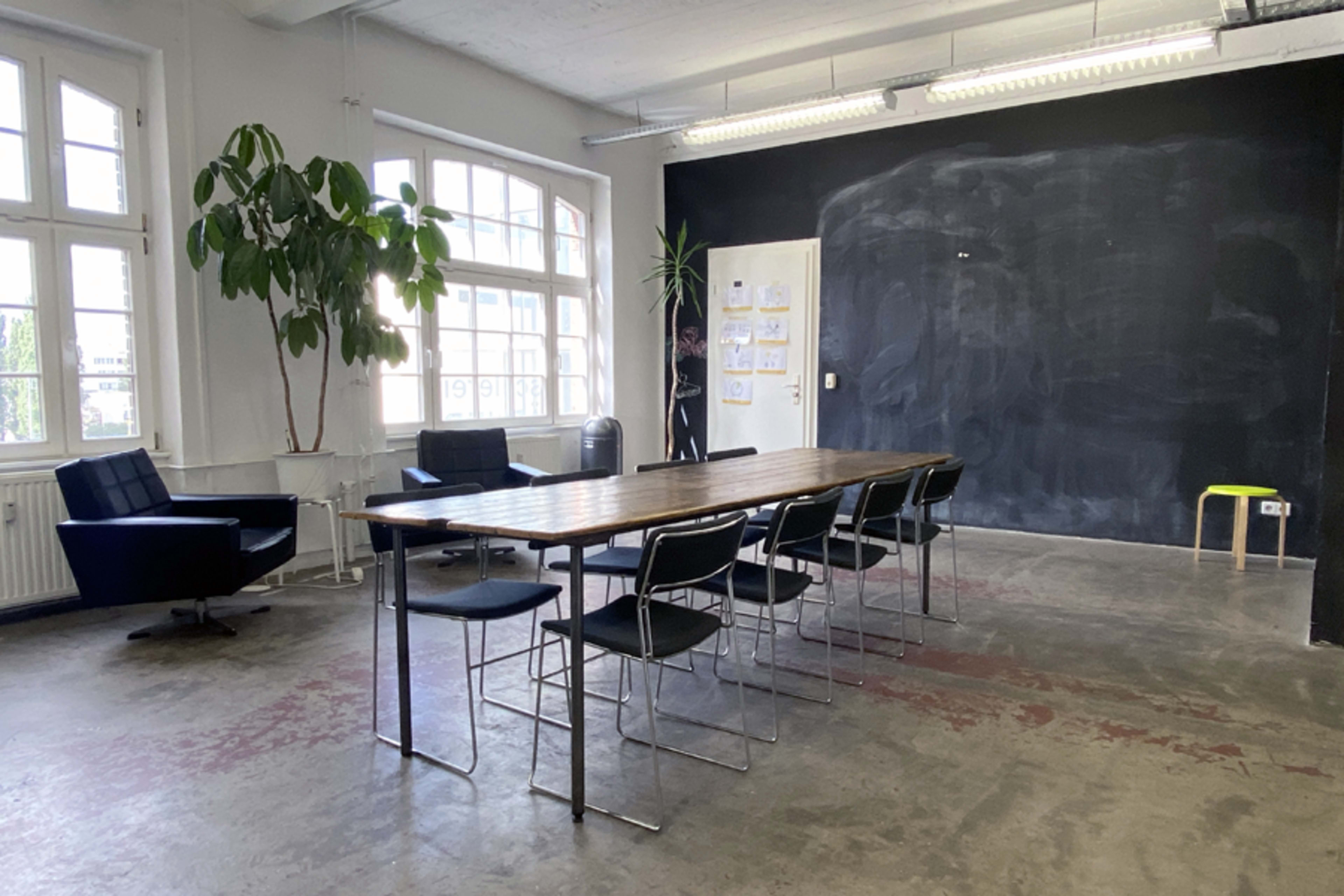 A spacious meeting room featuring a long wooden table surrounded by black chairs, with large windows and a chalkboard wall.
