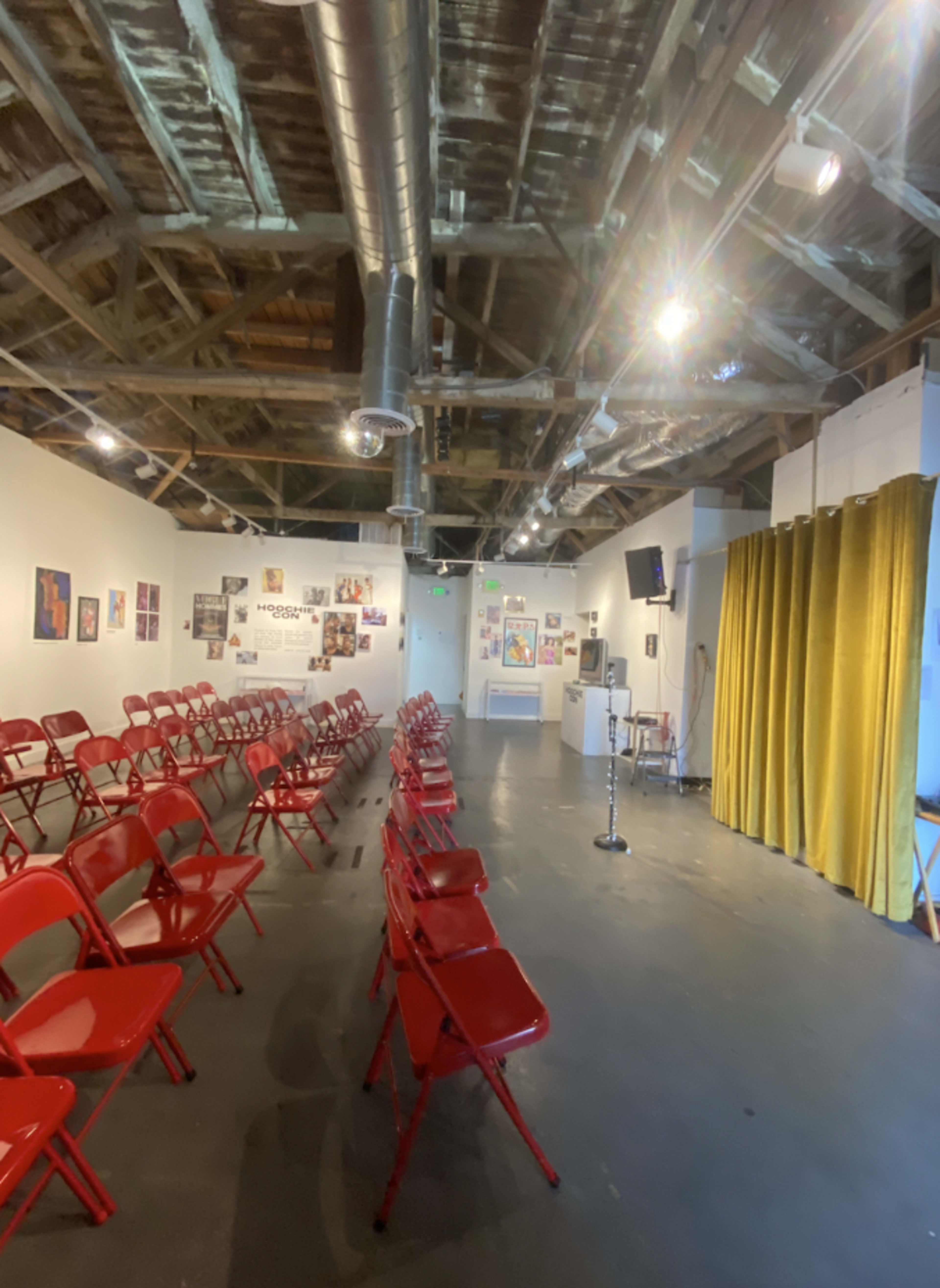 A spacious room with red folding chairs arranged in rows, a stage draped with yellow curtains, and various posters displayed on the walls.