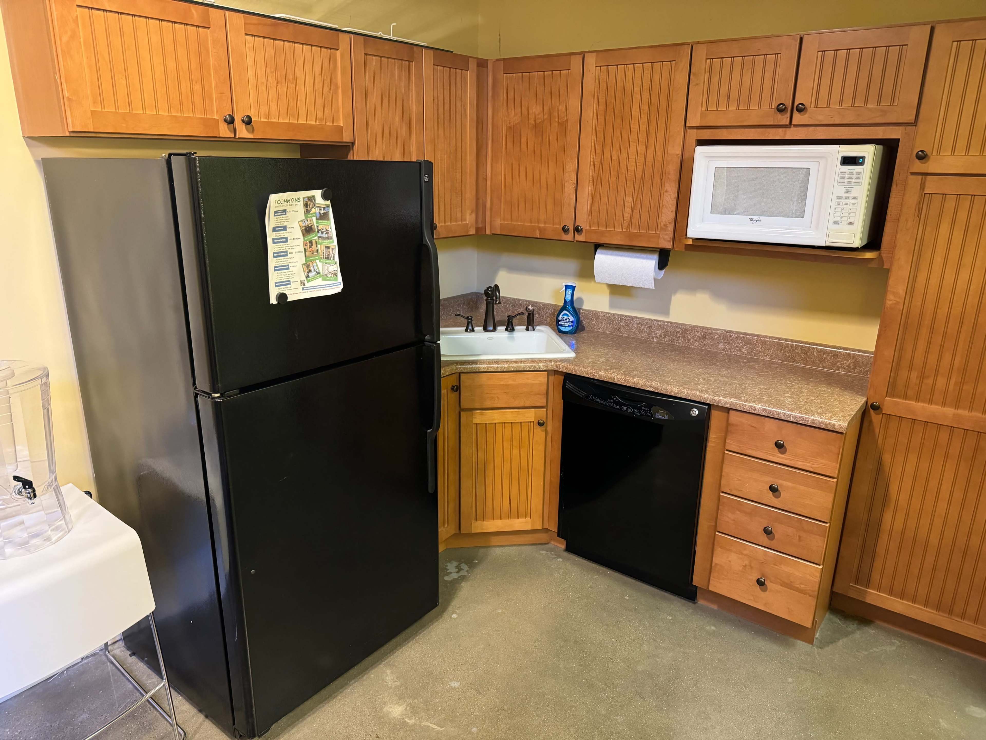The image shows a kitchen area featuring wooden cabinets, a black refrigerator, a black dishwasher, a microwave on the counter, and a sink.