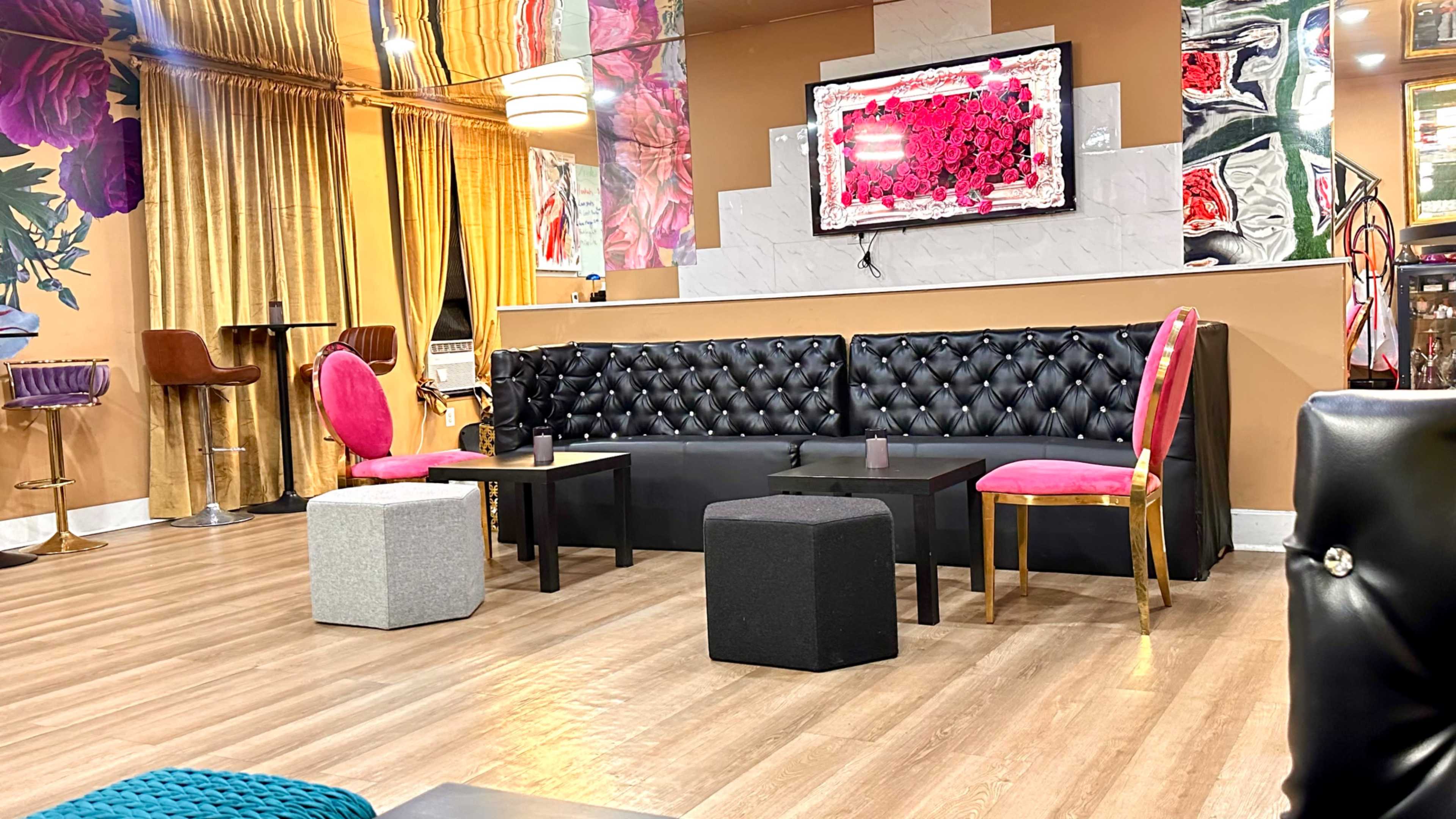 The image shows a modern lounge with a black tufted sofa, colorful pink chairs, and small coffee tables arranged on wooden flooring, surrounded by decorative floral and abstract wall art.