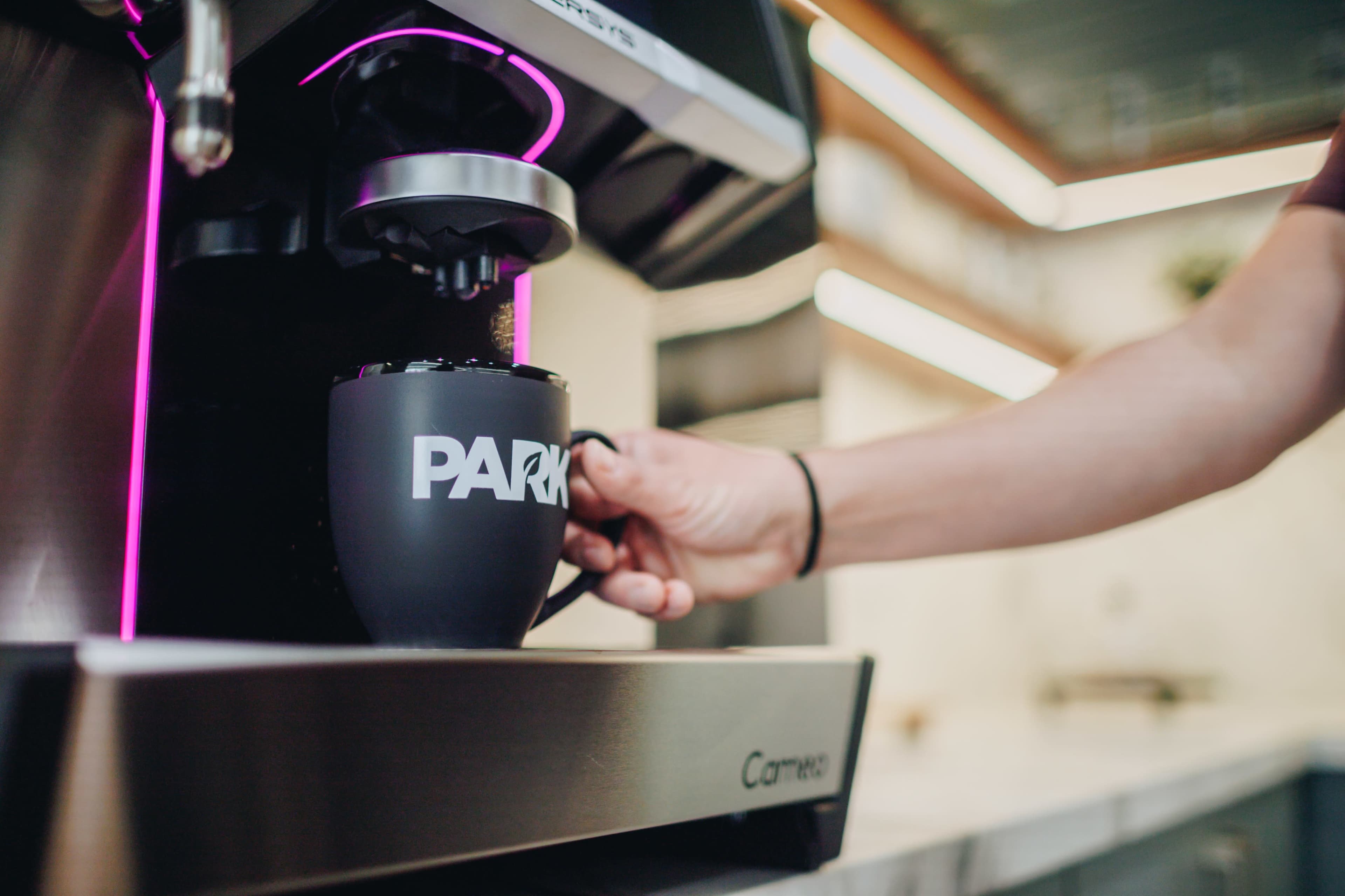 A person is pouring coffee from a machine into a black mug with the word "PARK" printed on it.
