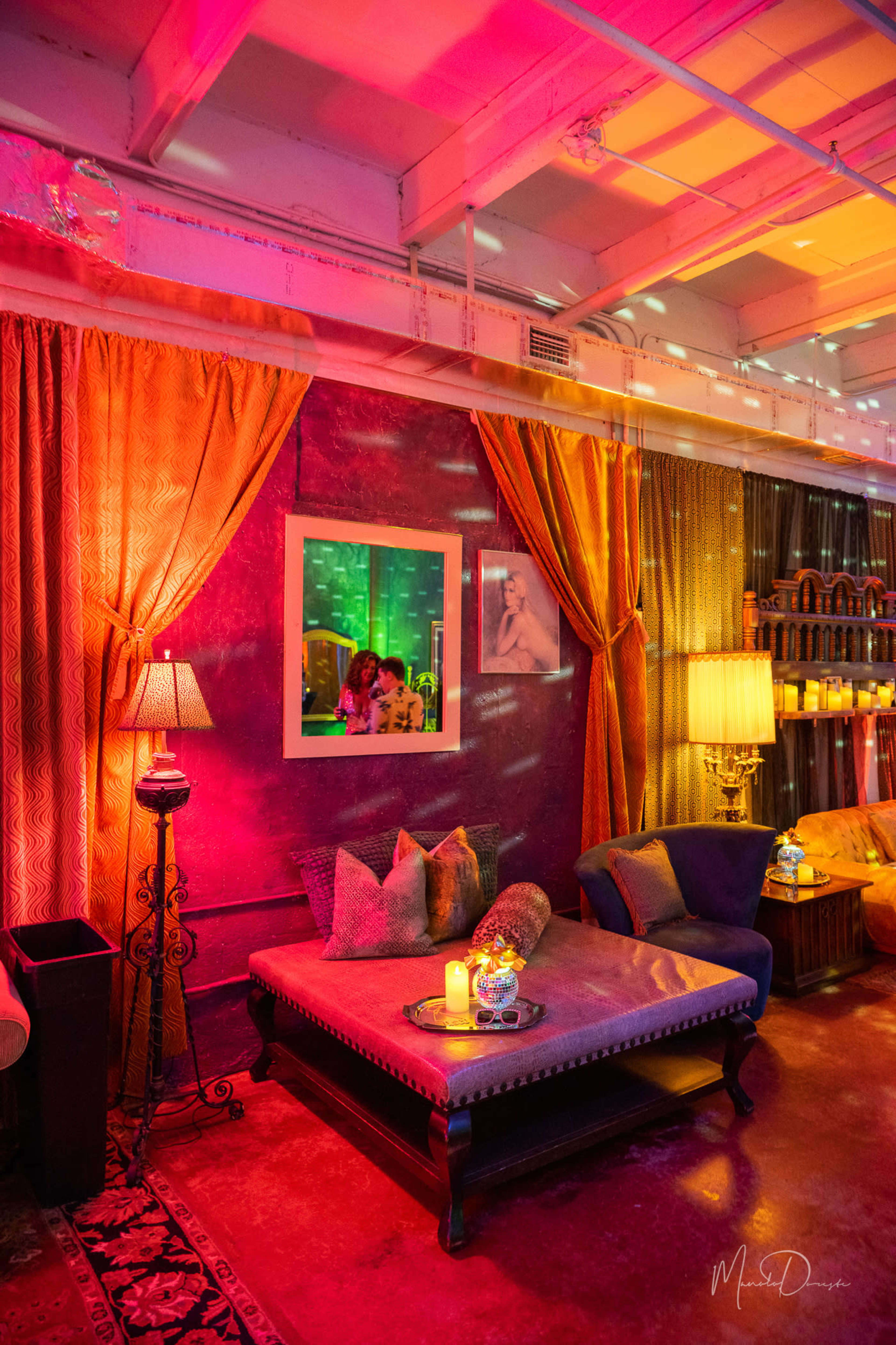 A softly lit lounge area with colorful lighting, decorative curtains, and a seating arrangement featuring a coffee table and lamps.