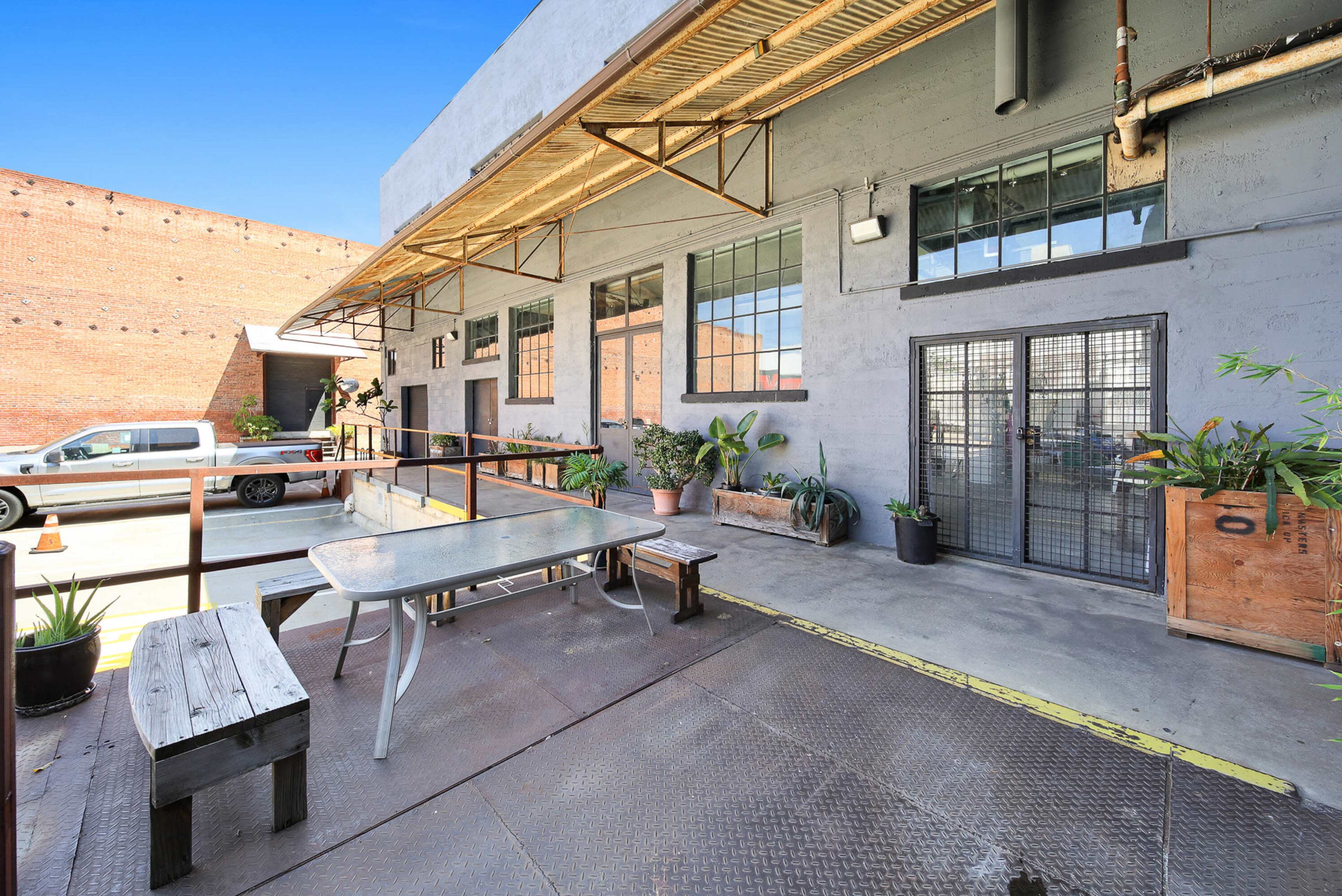 The image shows an industrial-style outdoor seating area with a metal table, wooden benches, and several potted plants, adjacent to a large building with multiple windows.
