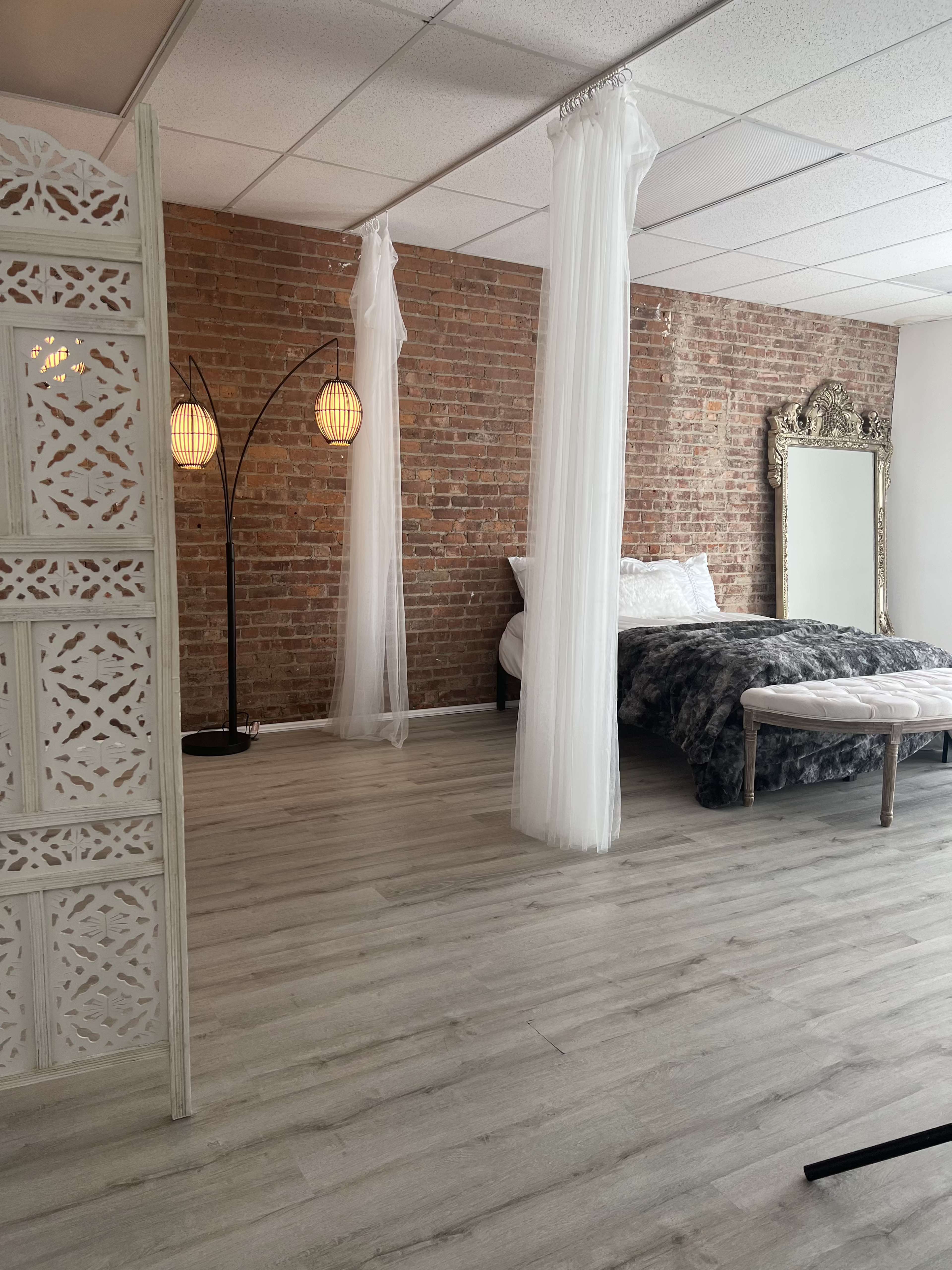 A bedroom with exposed brick walls, a bed draped in gray bedding, sheer white curtains hanging from the ceiling, and a decorative mirror beside a modern floor lamp.