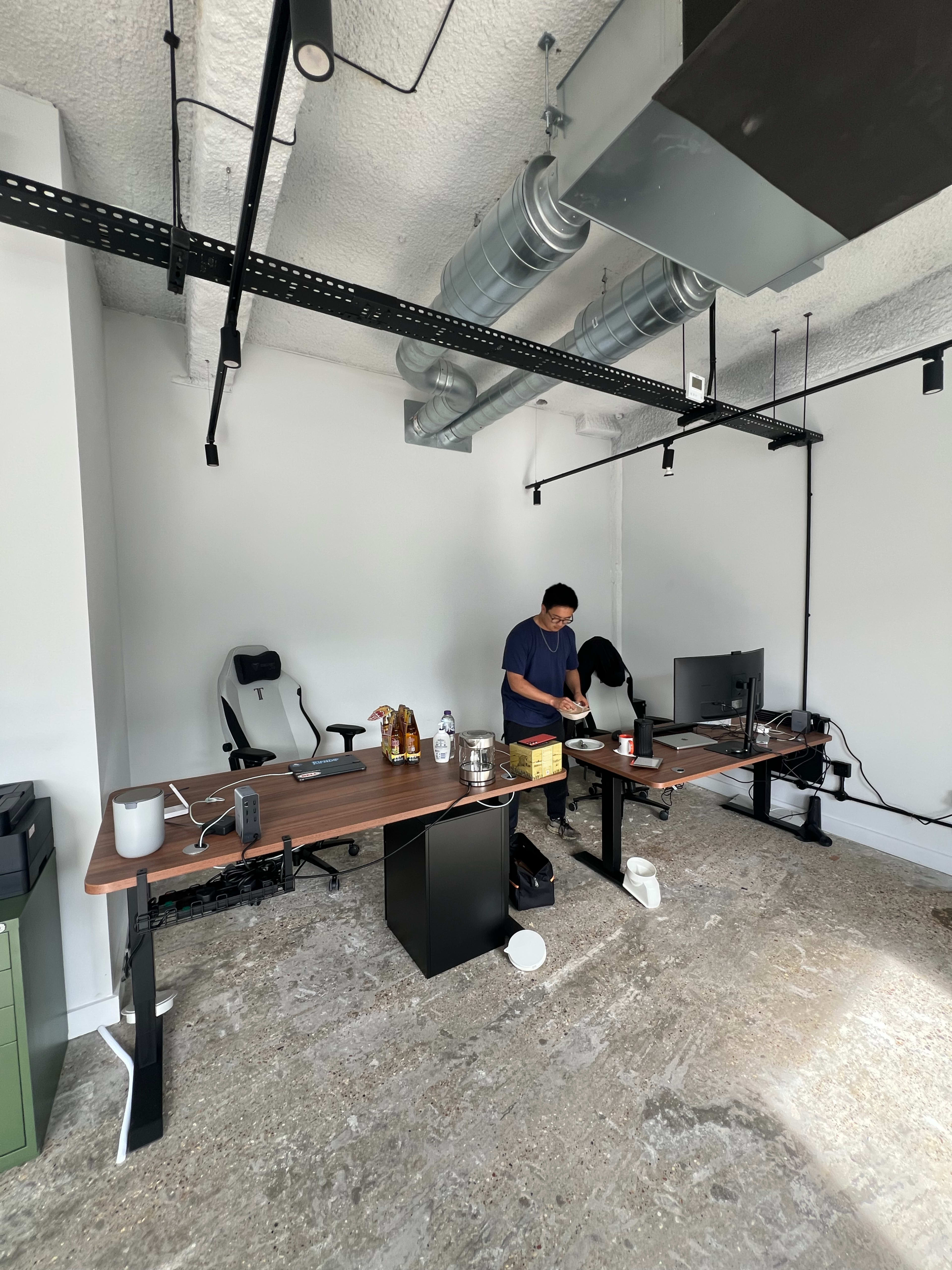 A person is standing at a desk in a sparsely furnished office space, with two empty ergonomic chairs and a computer monitor on the desk.