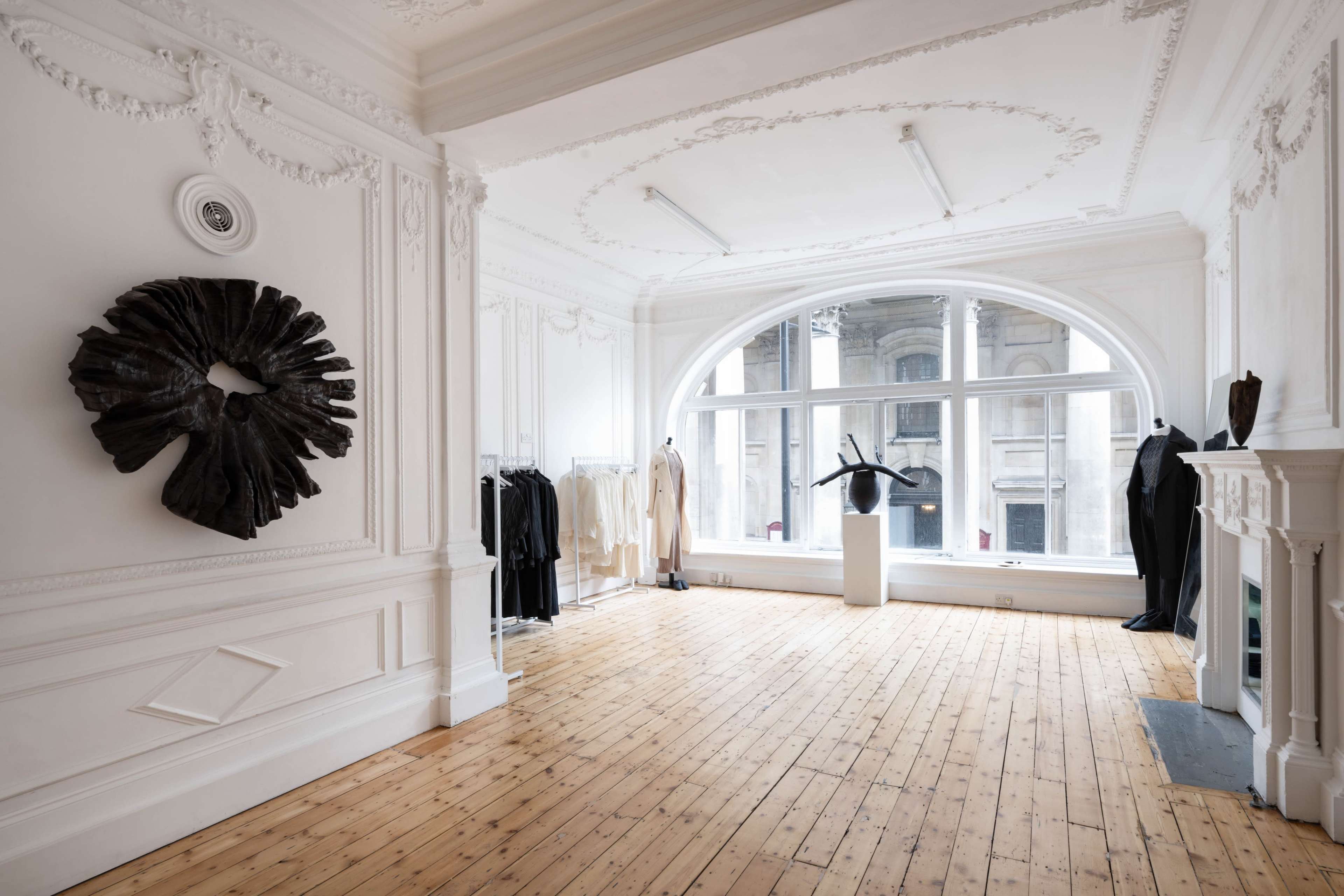 A spacious, elegantly designed clothing store featuring a large window, white walls, and wood flooring, with minimalist clothing displays and modern art pieces.