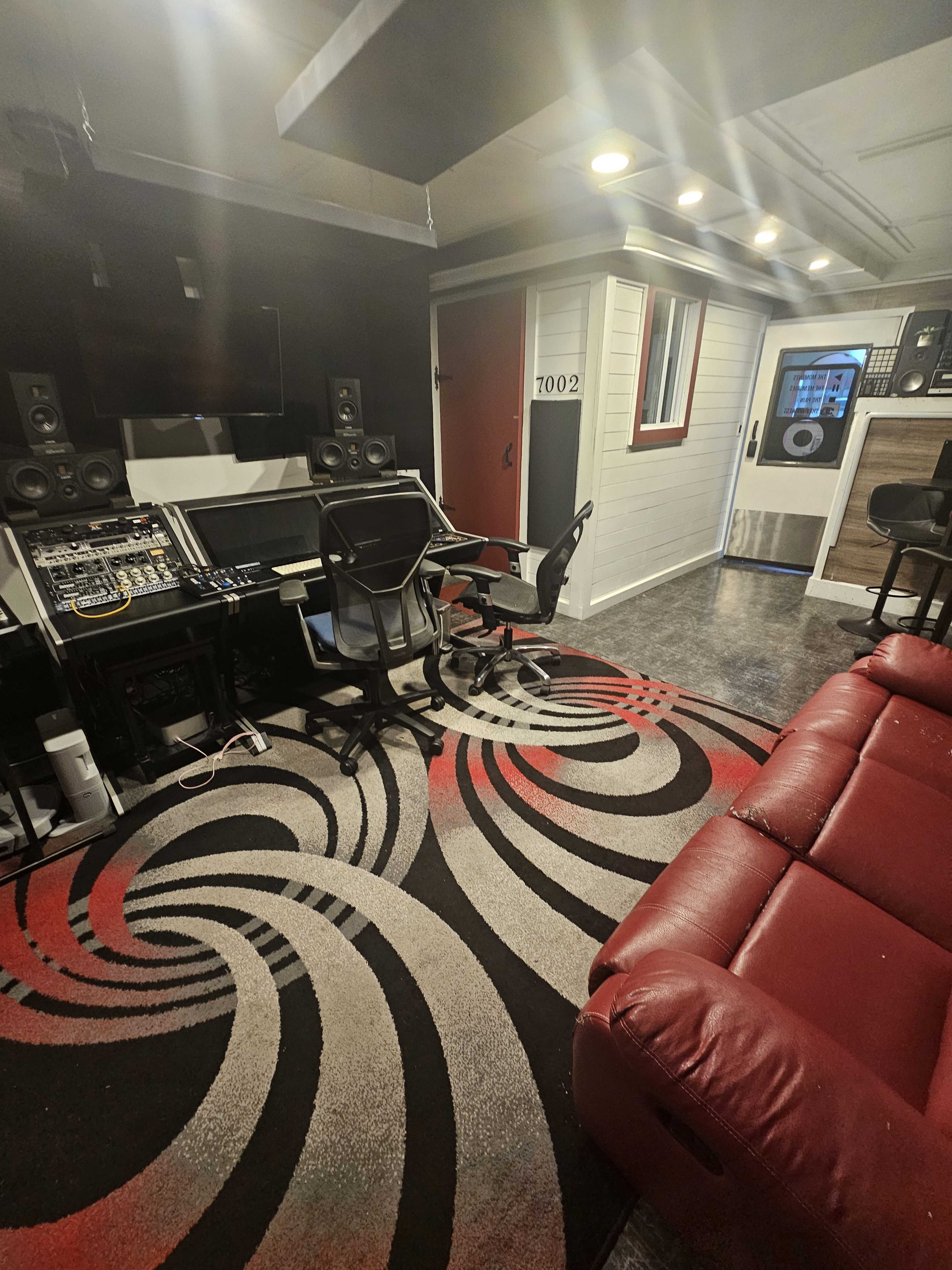 The image shows a small music production studio featuring a control desk with audio equipment, two chairs, a red leather couch, and a patterned rug on the floor.