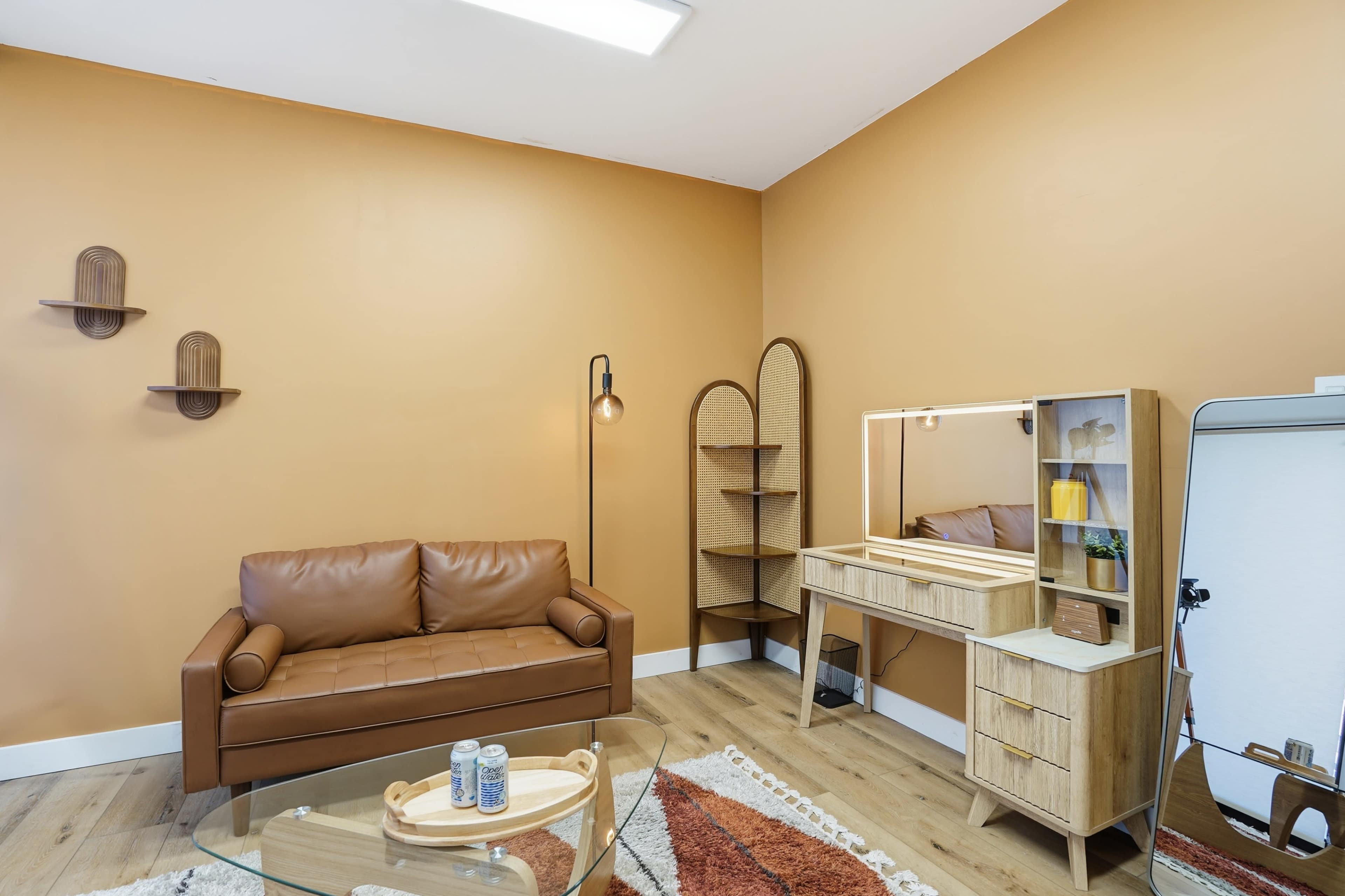 The image shows a cozy room with an orange wall, a brown leather sofa, a glass coffee table, and a wooden vanity with a mirror and decorative items.