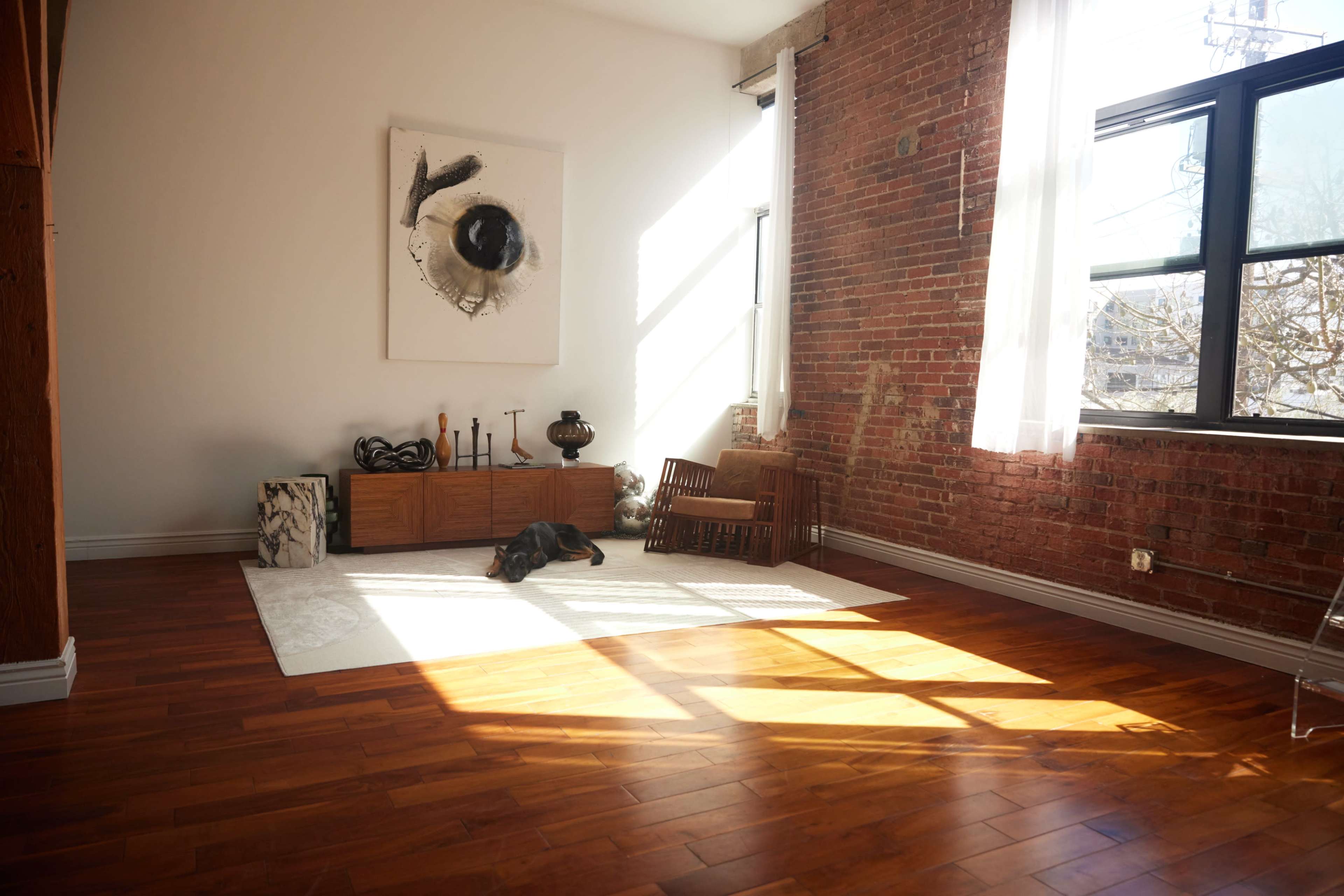 A spacious room features exposed brick walls, wooden flooring, and a large window casting shadows on a white area rug.