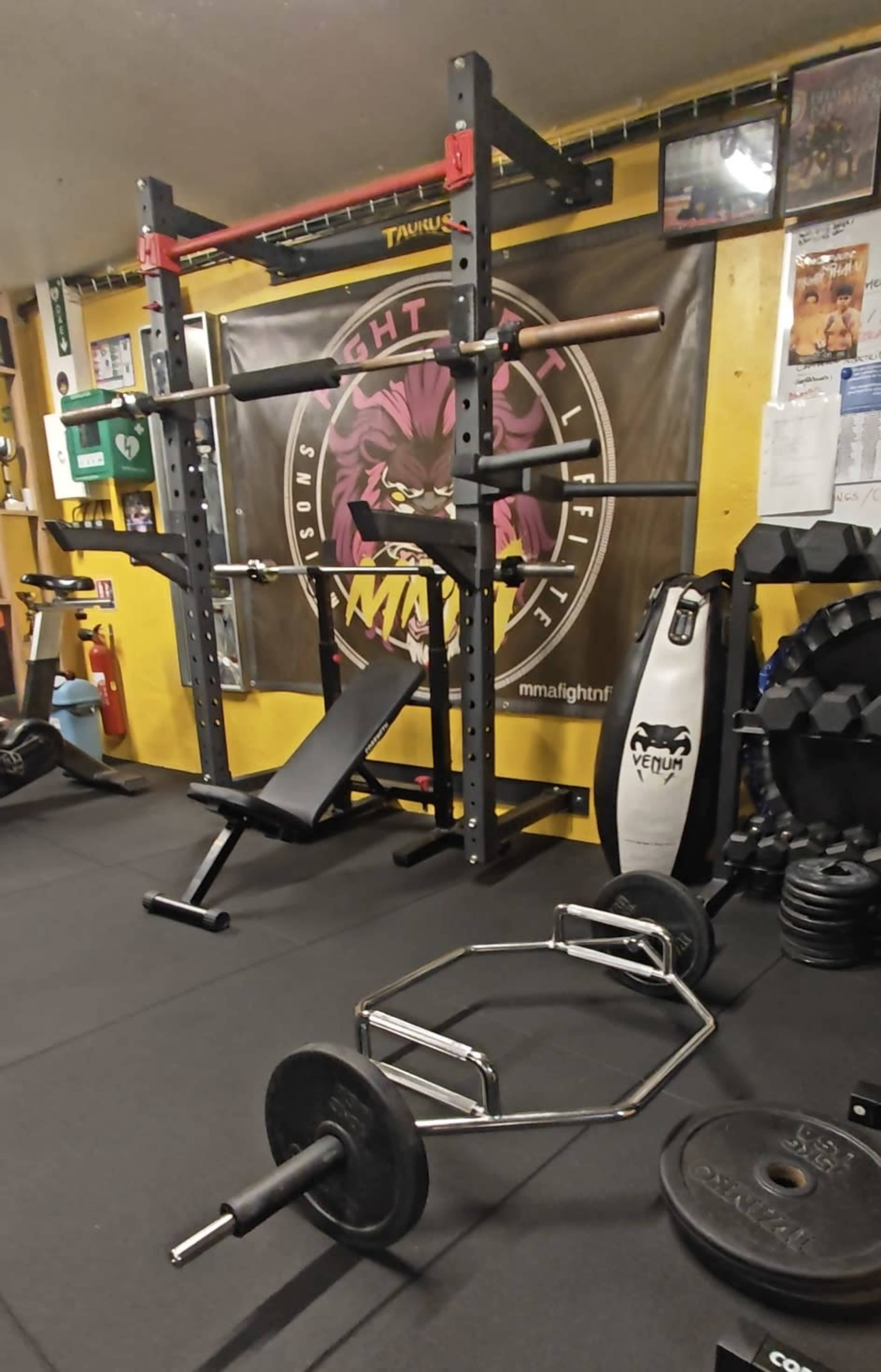 The image shows a gym setup featuring a squat rack, a weight bench, a barbell, a hex bar, and a variety of weights on a black mat.