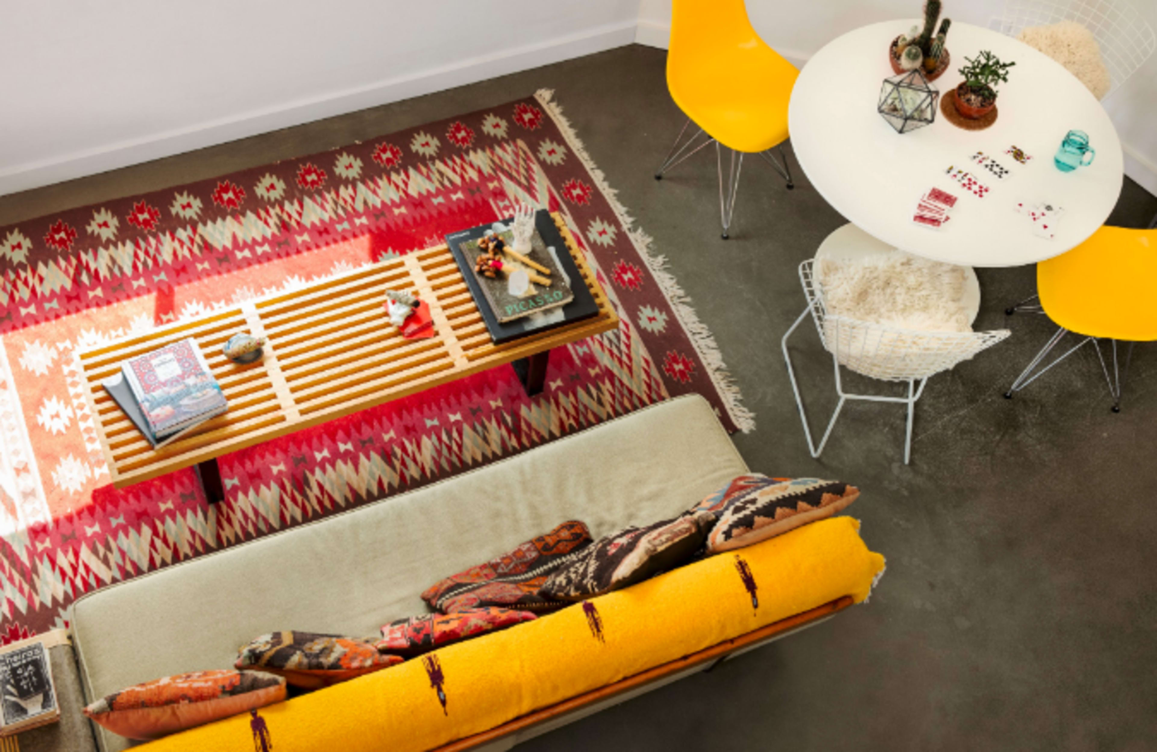 A brightly decorated living space features a yellow sofa with patterned cushions, a wooden coffee table, a round dining table with yellow chairs, and a colorful rug.