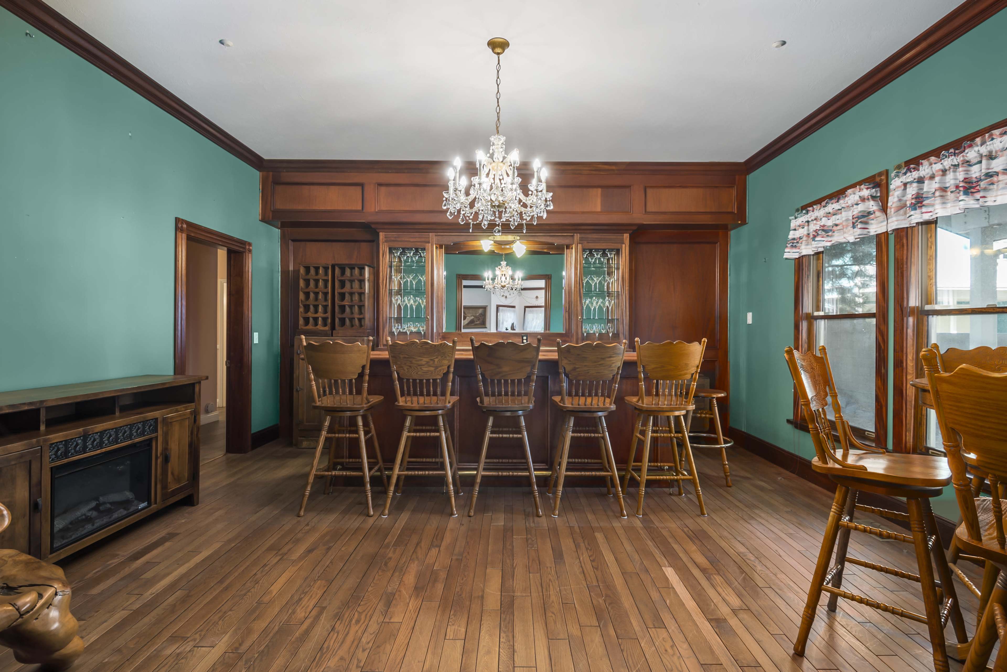 A spacious bar area features a chandelier, wooden bar stools, and a wine rack, all surrounded by teal walls.
