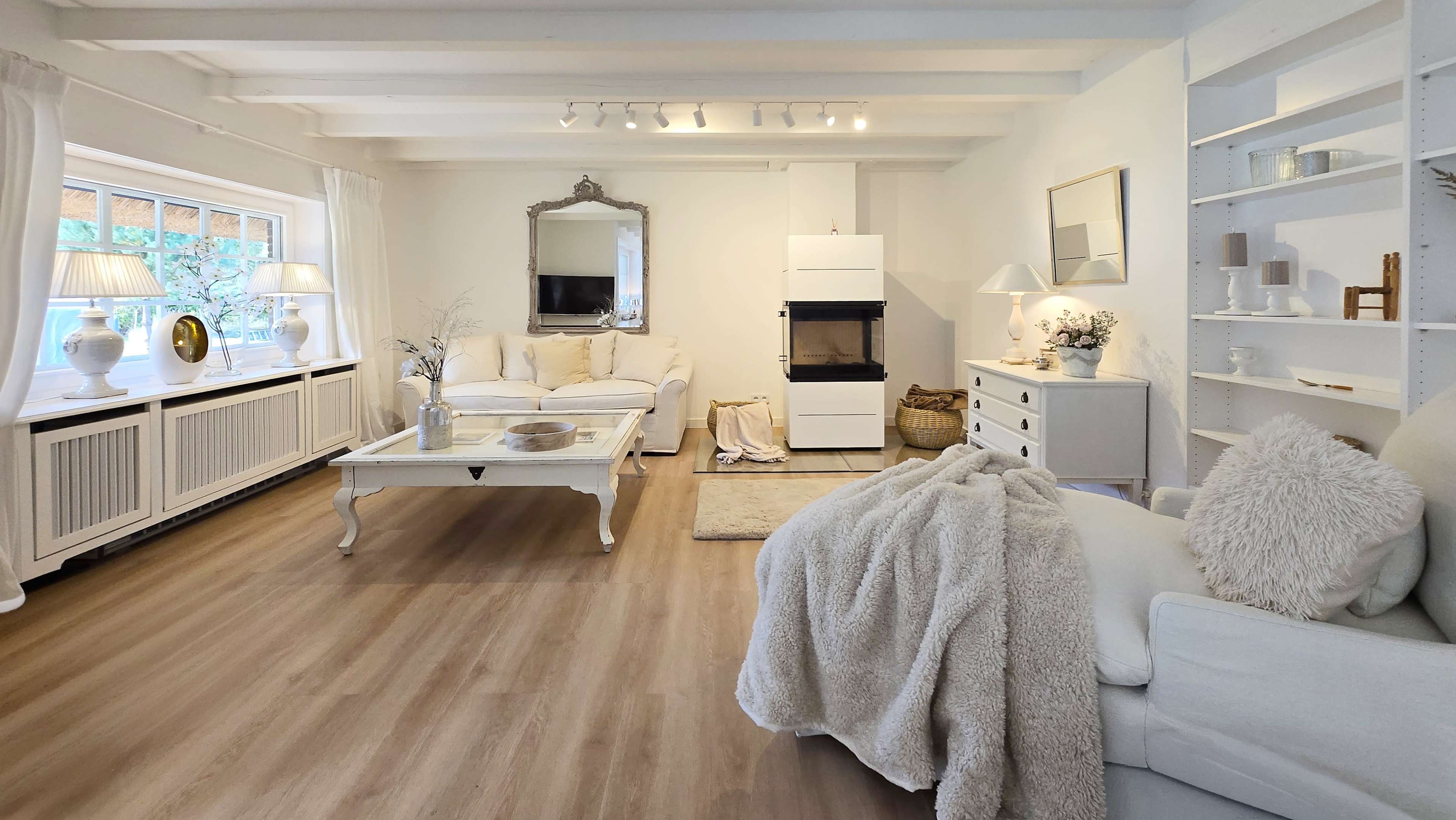 The image shows a bright living room with white furniture, a coffee table in the center, and large windows letting in natural light.