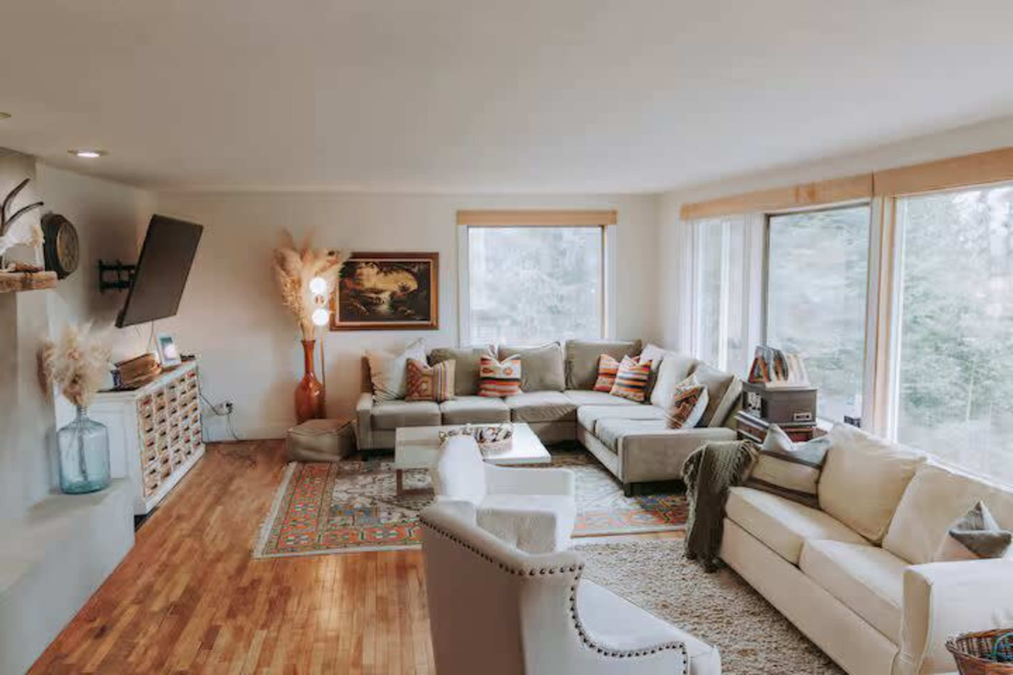 The living room features multiple seating areas with a light-colored sectional sofa, wooden flooring, large windows, and a mounted television on the wall.