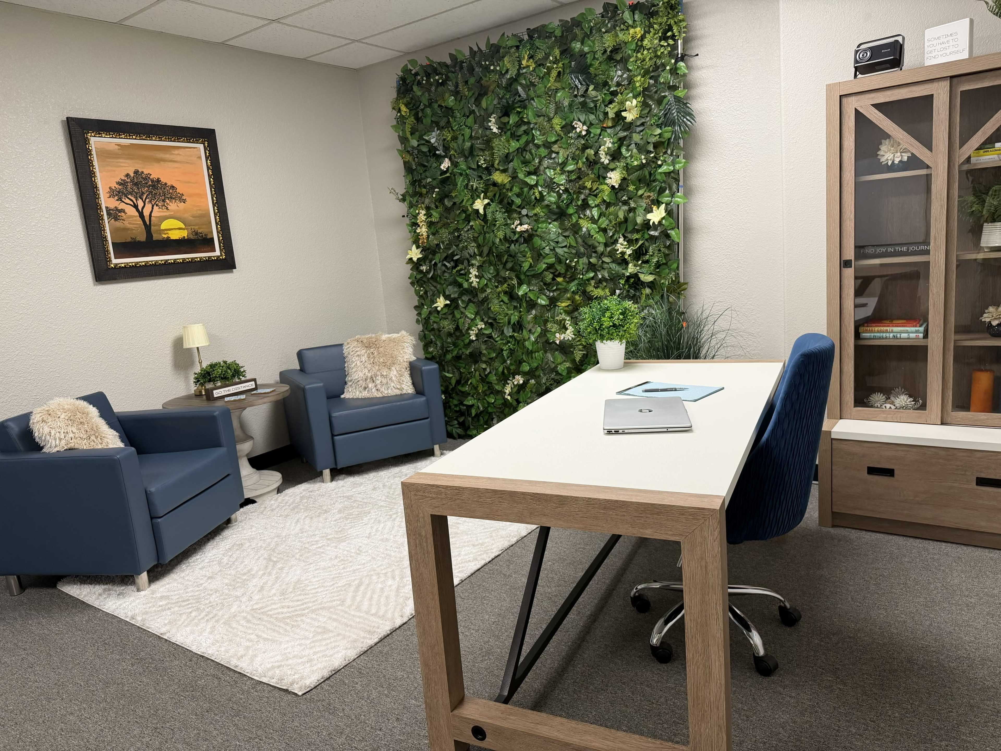 The image shows a modern office space featuring two blue armchairs, a large table with a chair, a decorative plant wall, and framed artwork on the walls.