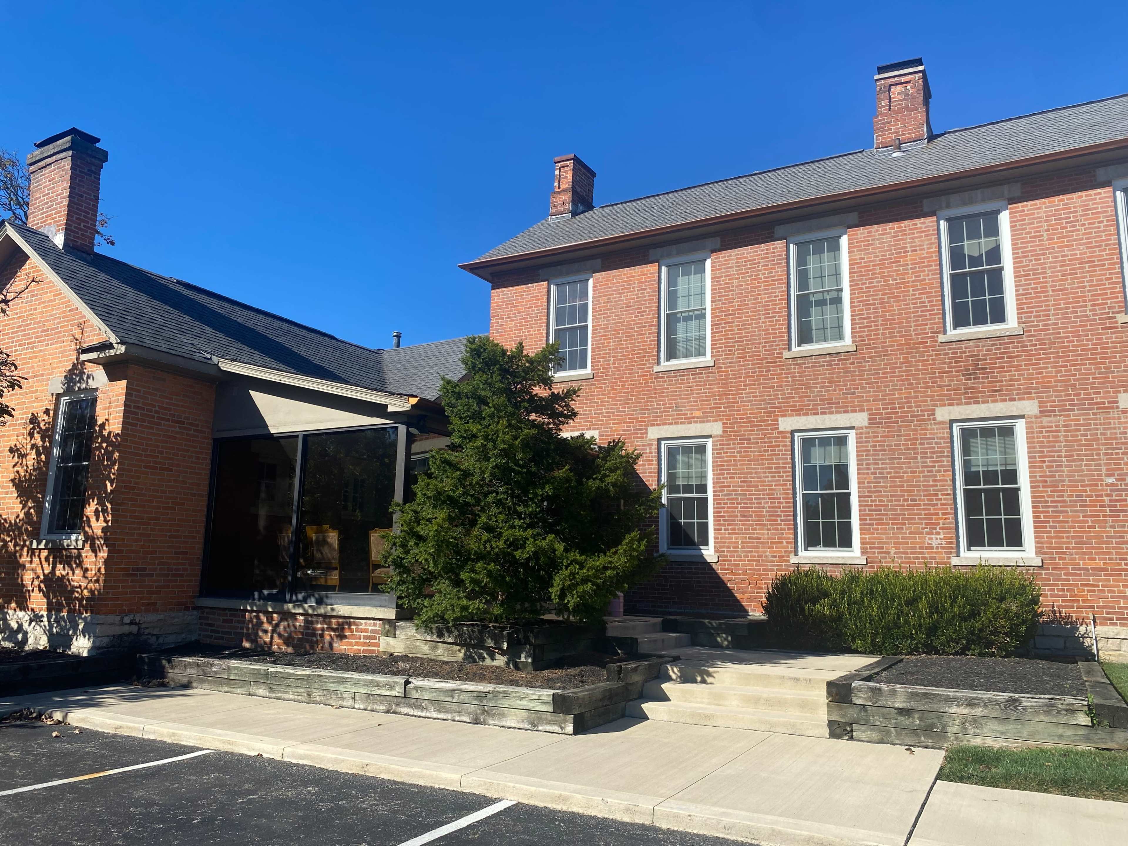 A brick building with two distinct sections, featuring large windows and a landscaped entrance, is situated under a clear blue sky.