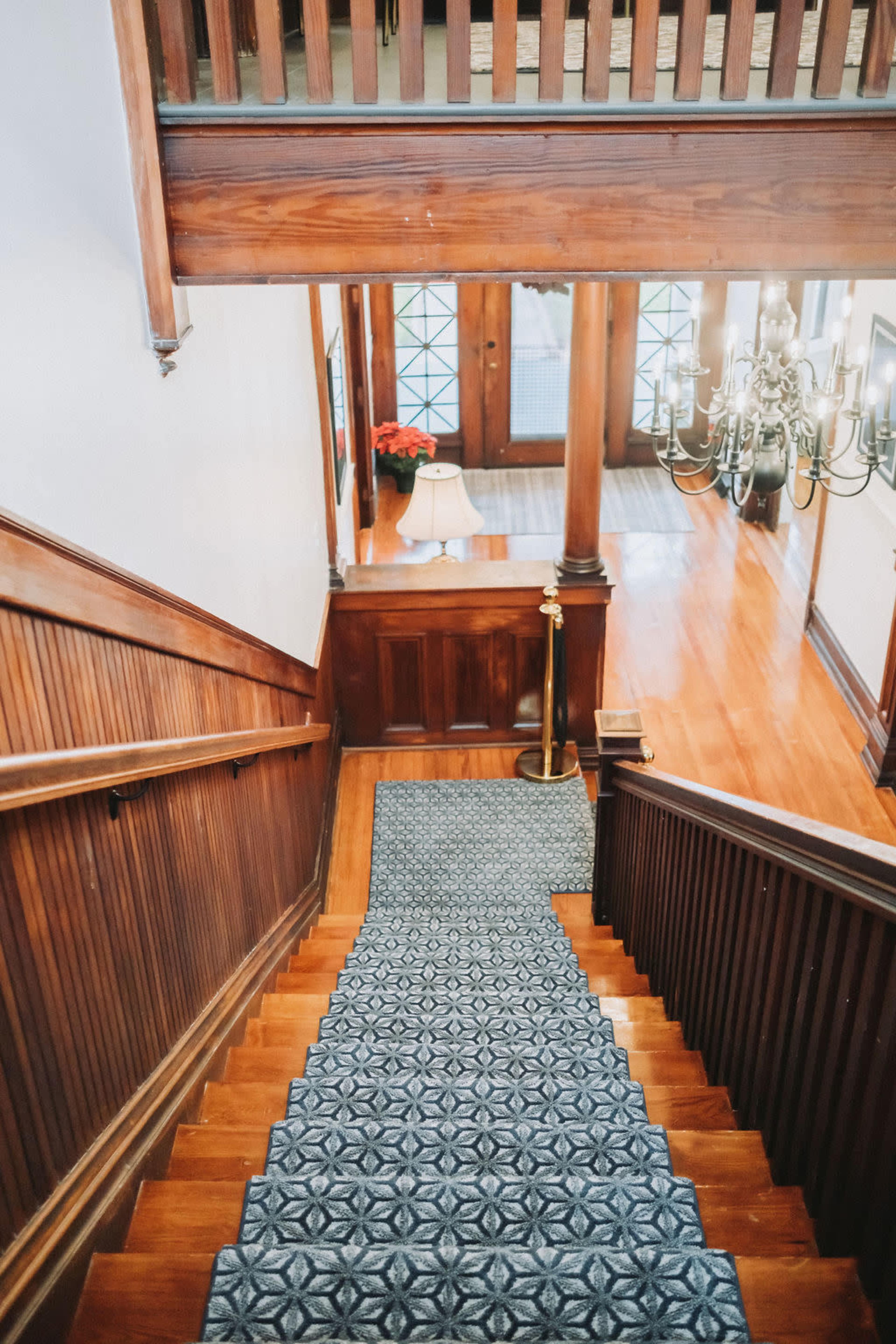 A wooden staircase leads down to a warmly lit foyer with a chandelier and a lamp on a side table.