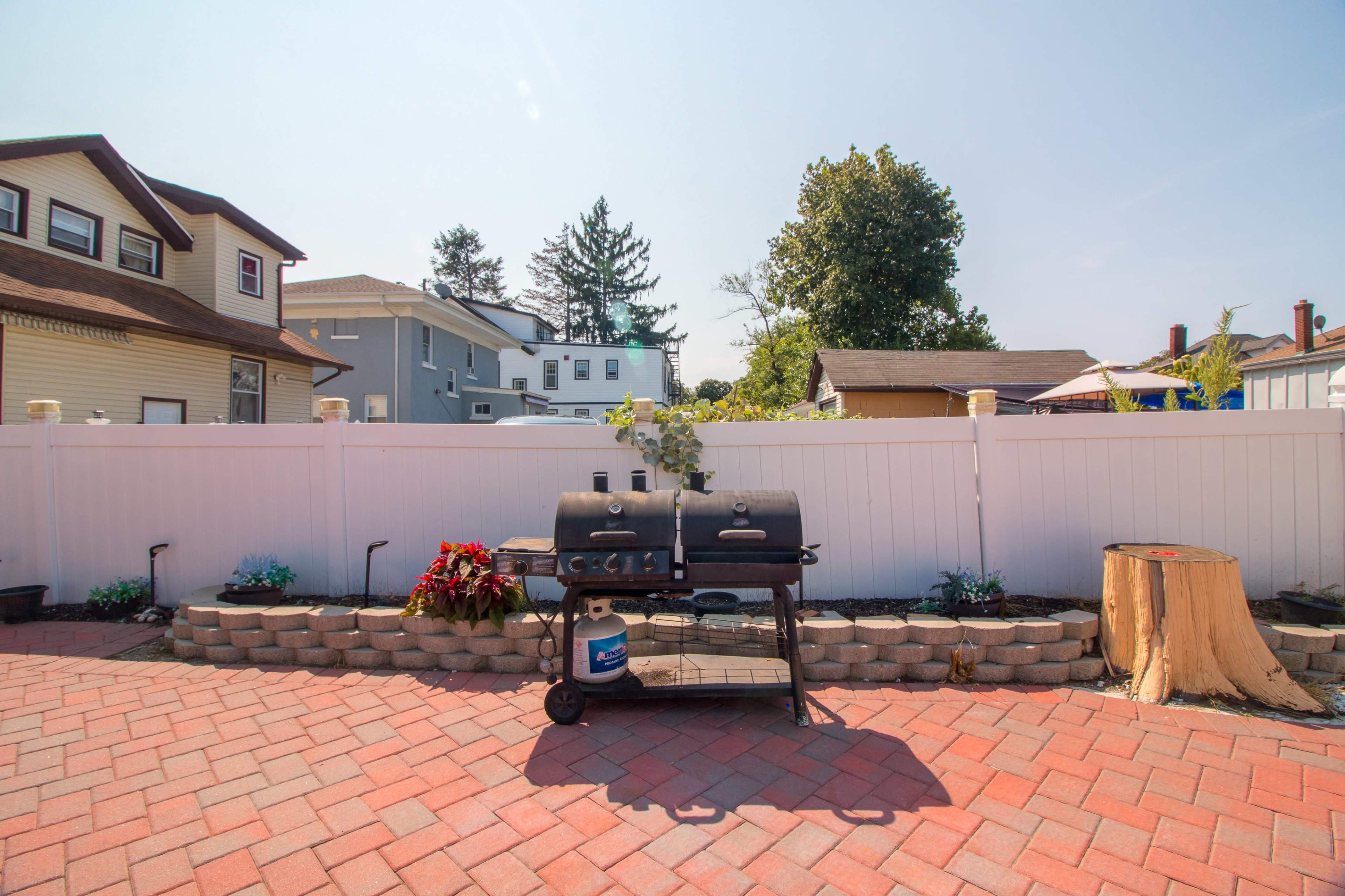A charcoal grill sits on a brick patio surrounded by a white fence and landscaped with plants and a tree stump.