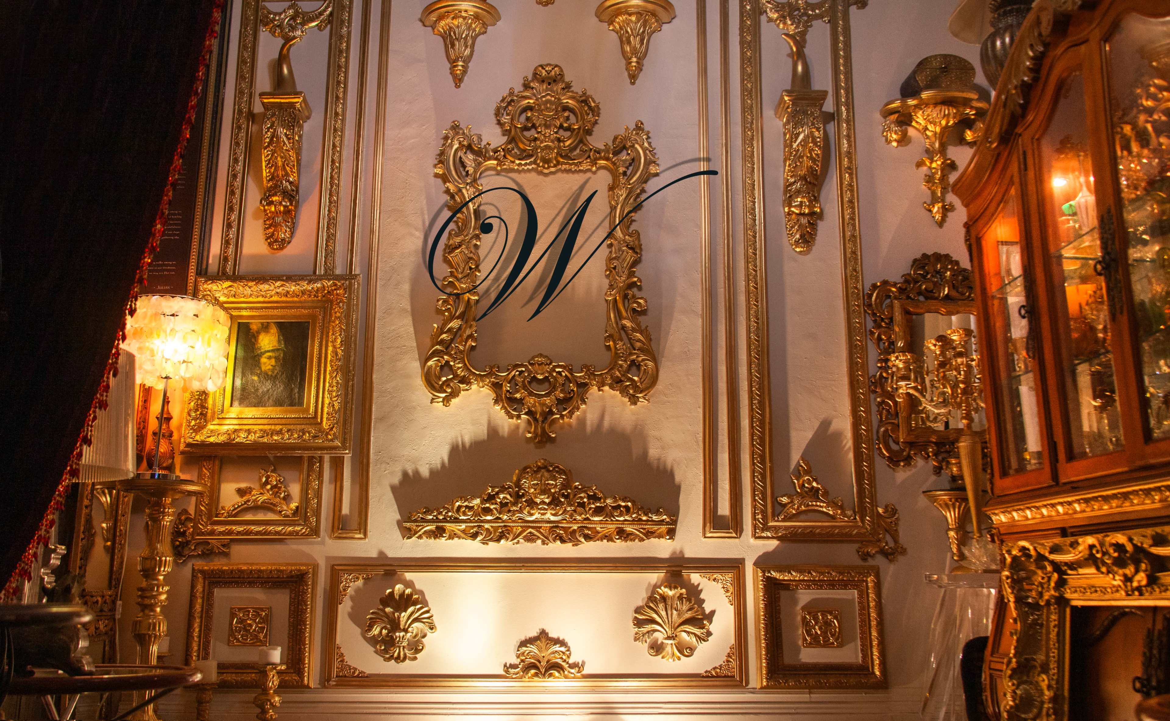 The image shows an ornate wall adorned with gold decorative elements and a large empty frame, with various antique items displayed nearby.