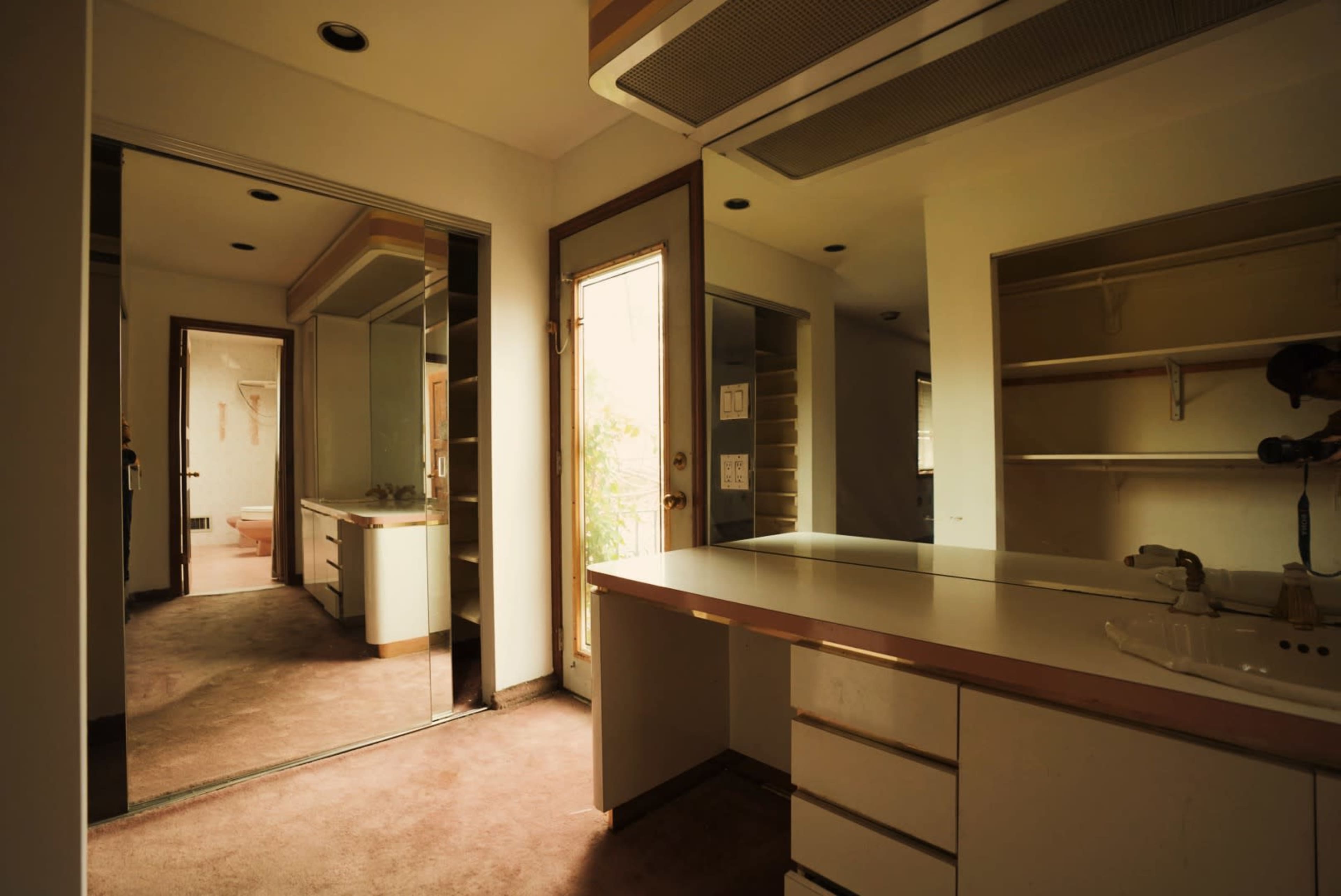 A bathroom features a vanity with a sink and mirror, adjacent to a door leading to an outdoor area, with shelves visible in the background.