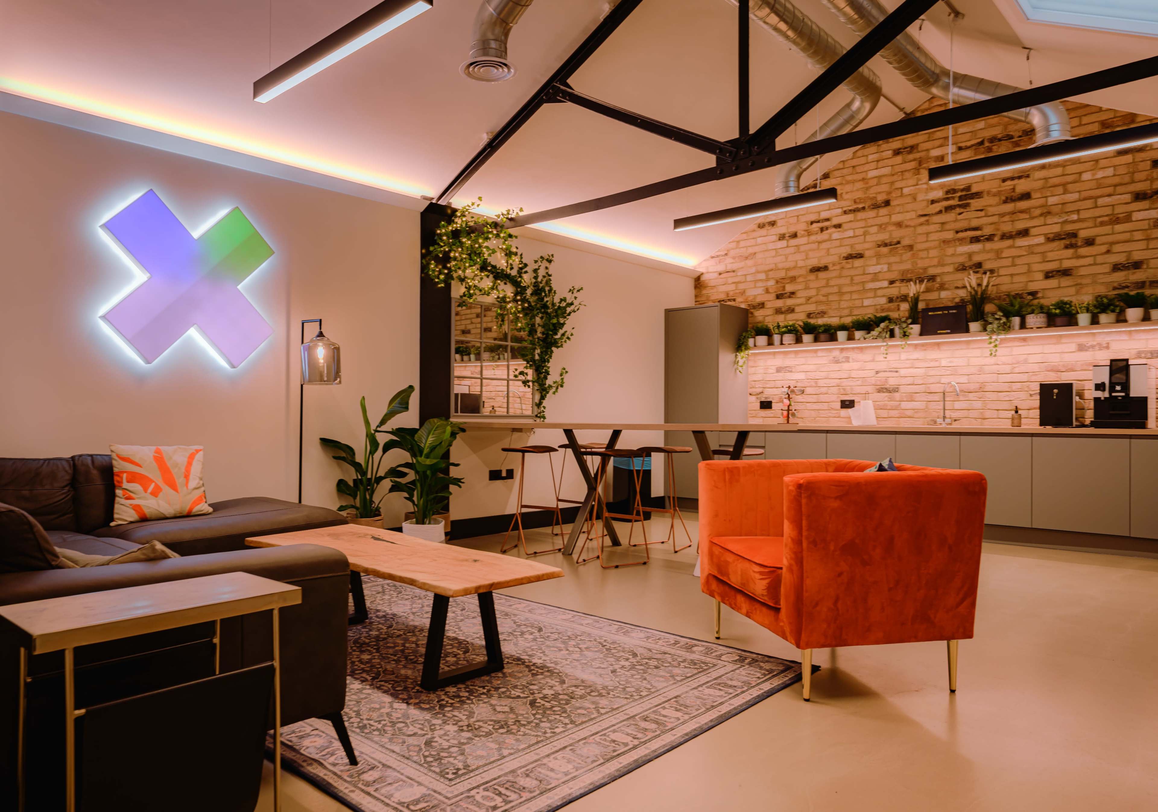 A modern, open-concept workspace featuring a lounge area with a dark sofa, an orange armchair, a wooden coffee table, and a kitchen with brick walls and greenery.