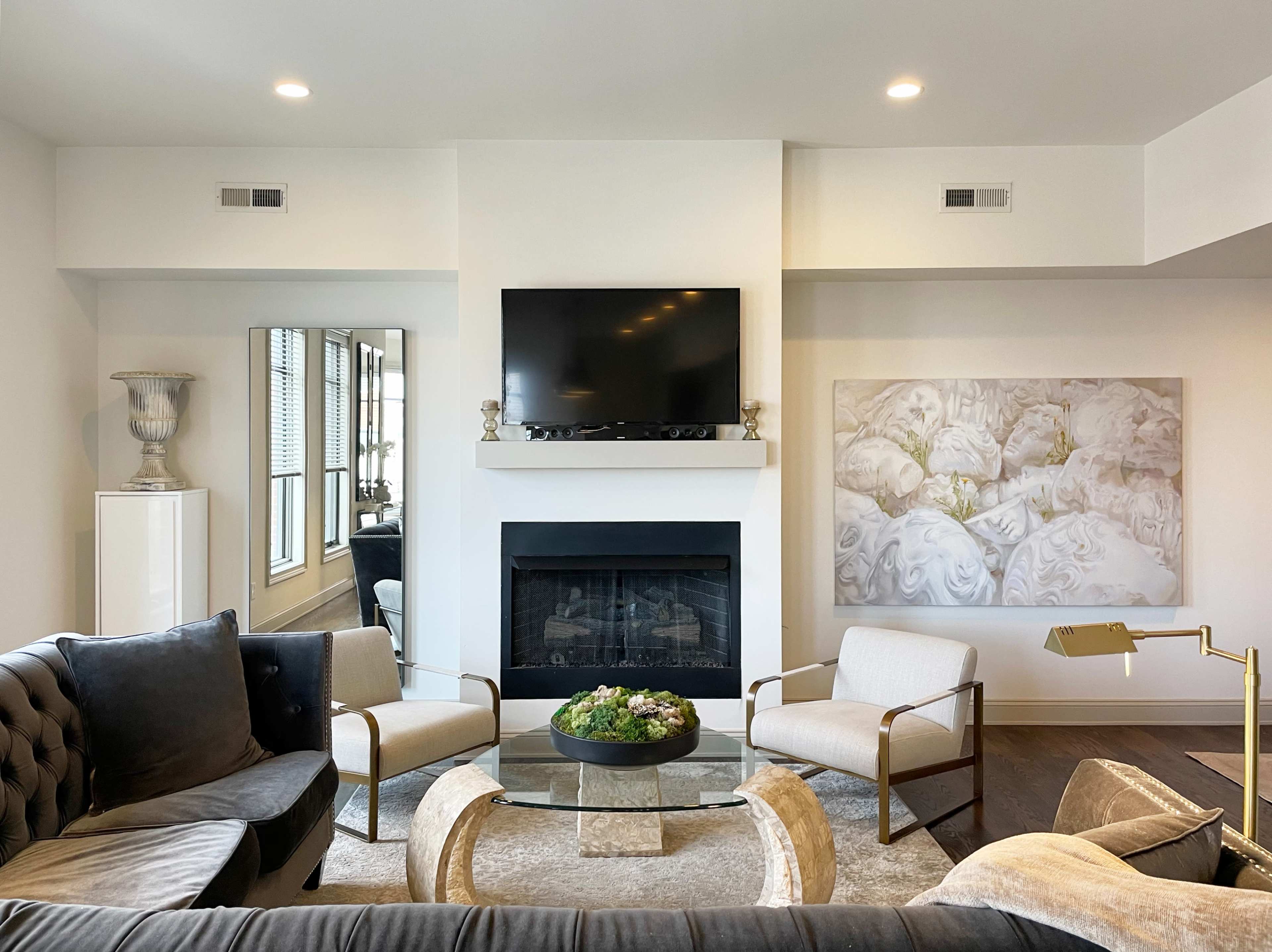 A modern living room features a fireplace, a wall-mounted TV, two armchairs, and a circular glass coffee table with a decorative centerpiece.