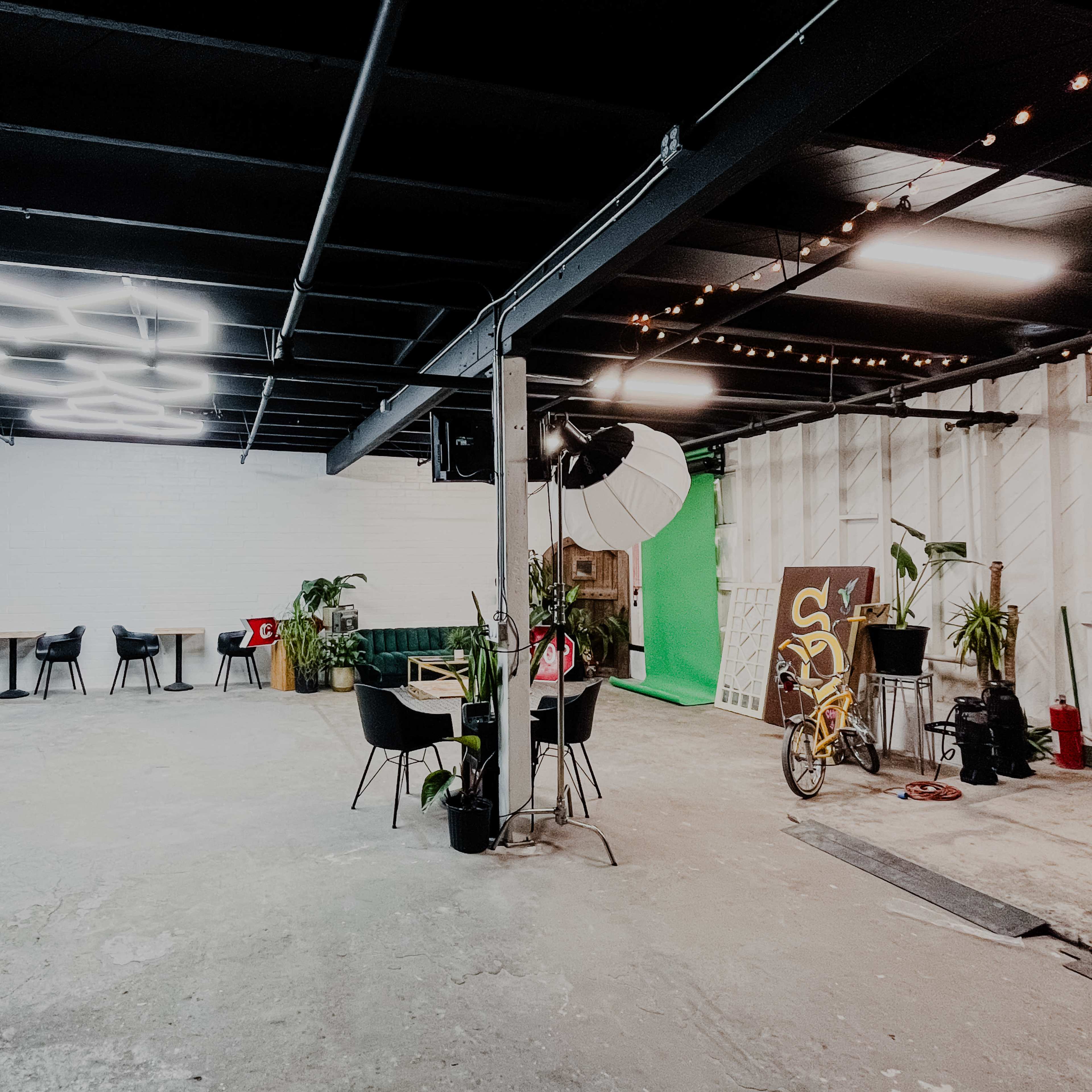 The image shows a spacious indoor area with a green backdrop, a table surrounded by chairs, and various plants scattered throughout.