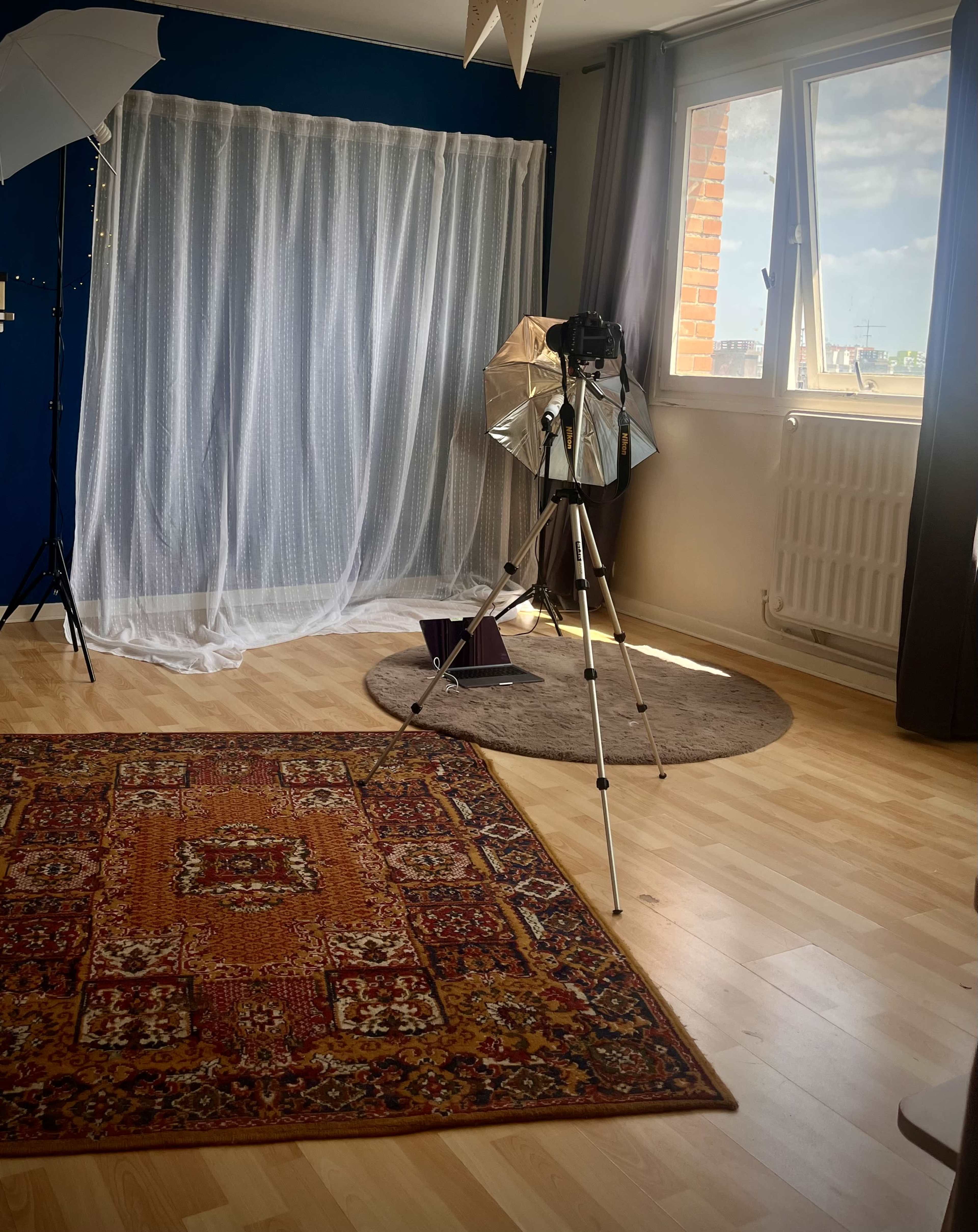 The image shows a photography studio with a backdrop curtain, a tripod with a camera, a light reflector, and a decorative rug on the wooden floor.