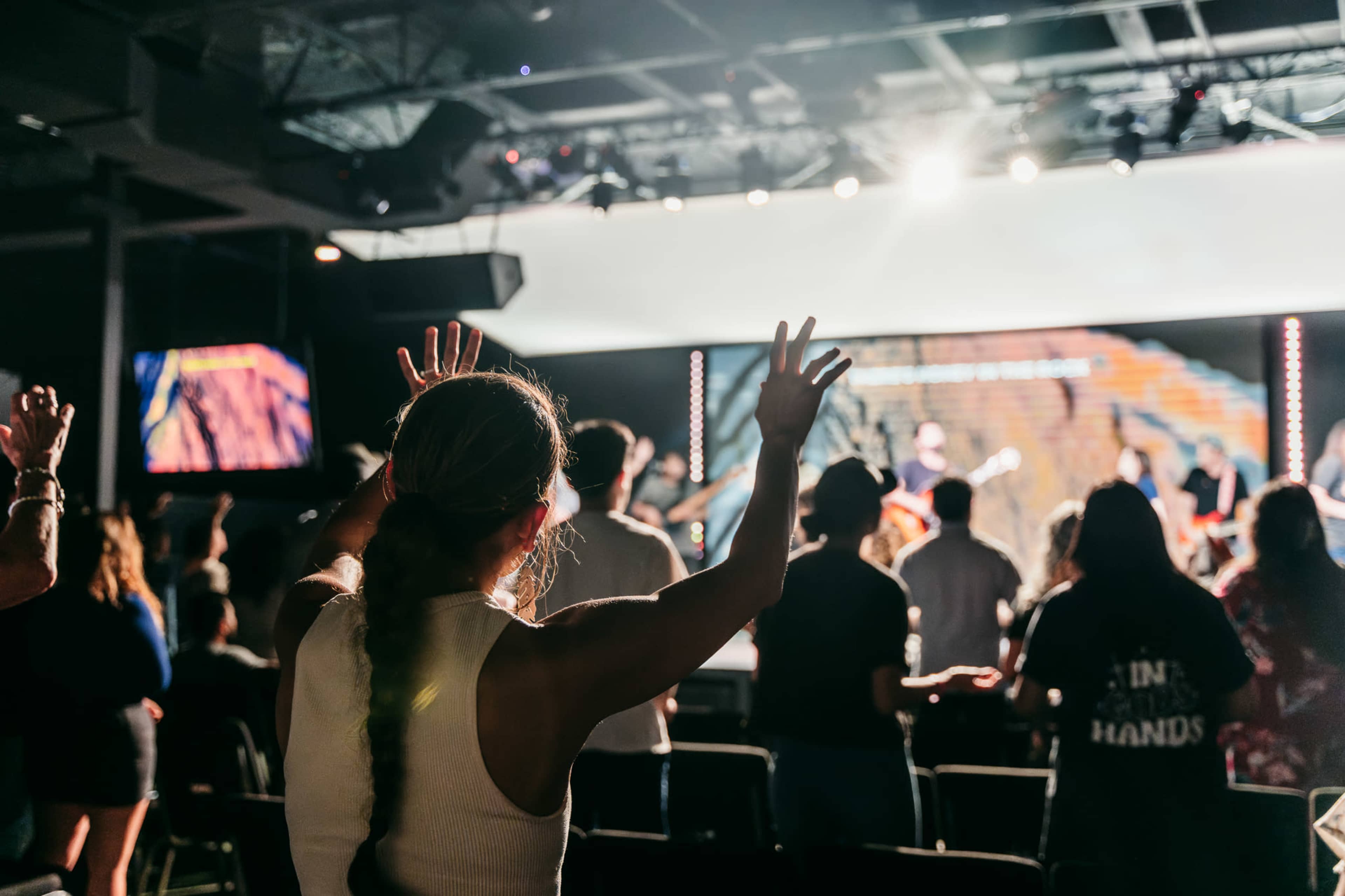 A group of people with raised hands worship during a live music performance in a dimly lit venue.