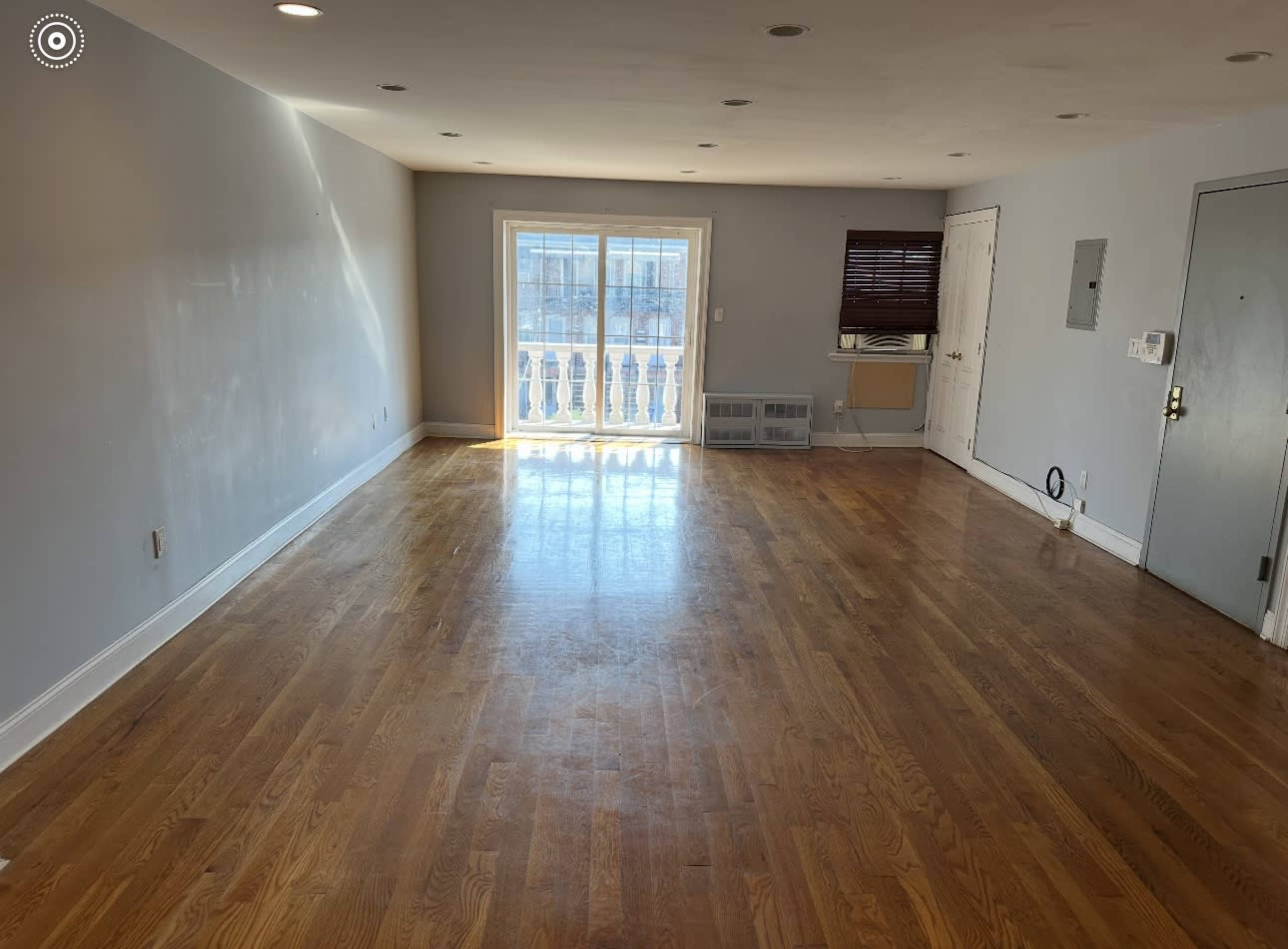 The image shows a spacious, empty room with hardwood flooring and a large window that provides natural light.