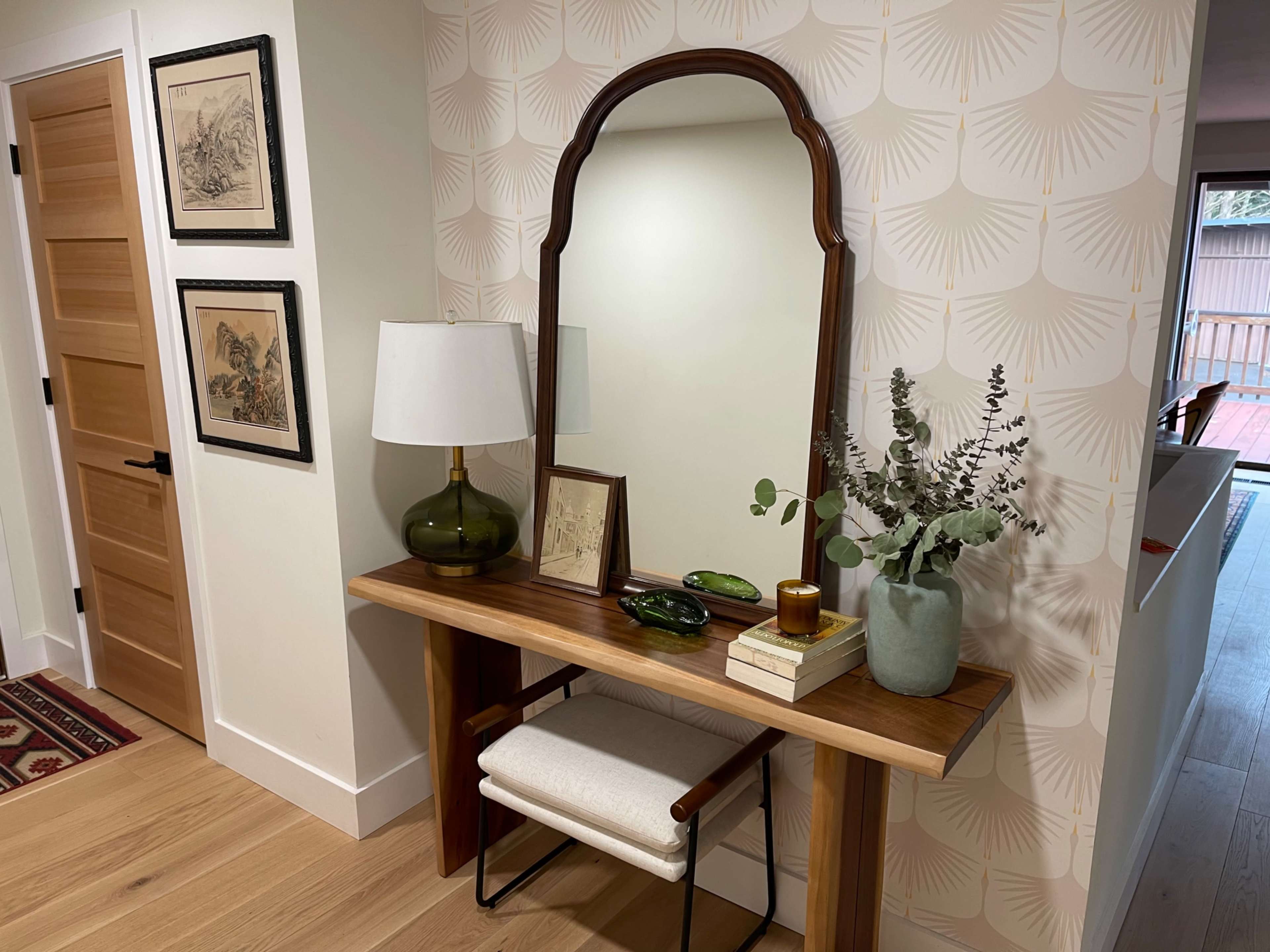 A wooden console table with a mirror above it is positioned against a patterned wall, accompanied by a lamp, decorative items, and a potted plant, while a doorway leads to another room.