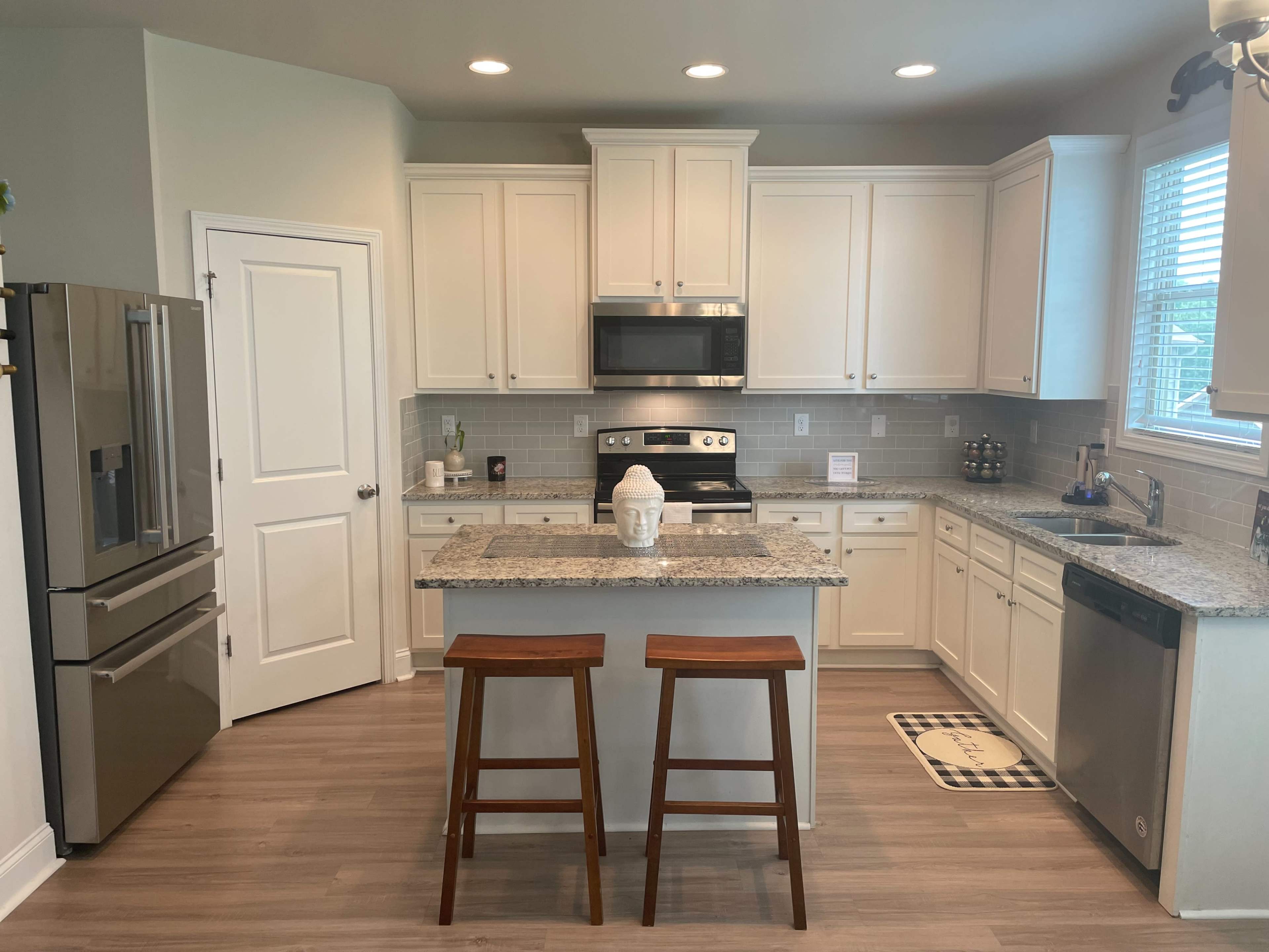 A modern kitchen features white cabinetry, granite countertops, stainless steel appliances, and two wooden stools at an island.