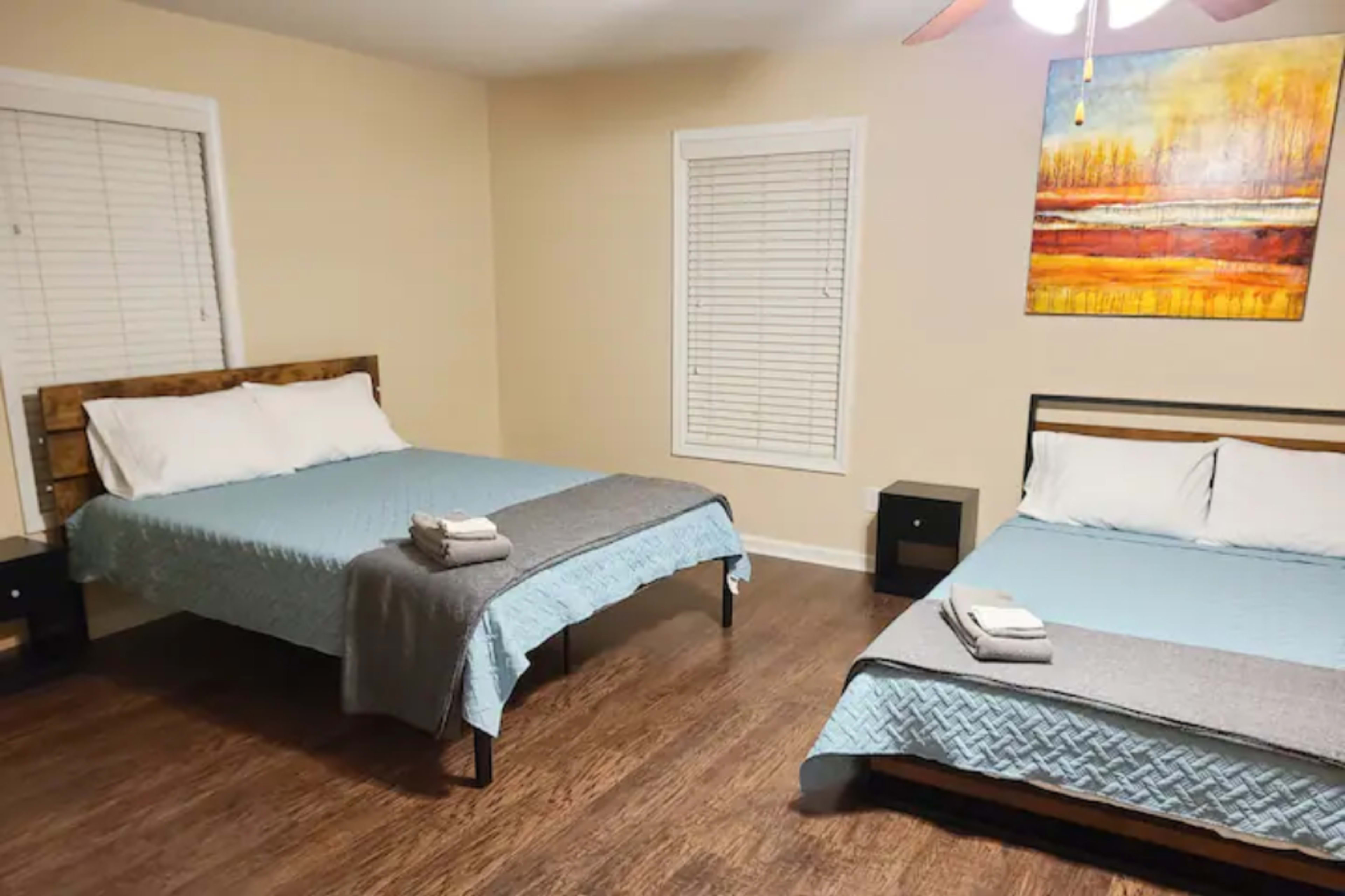 The image shows a bedroom featuring two beds with light blue covers, a wooden floor, and a colorful painting on the wall.