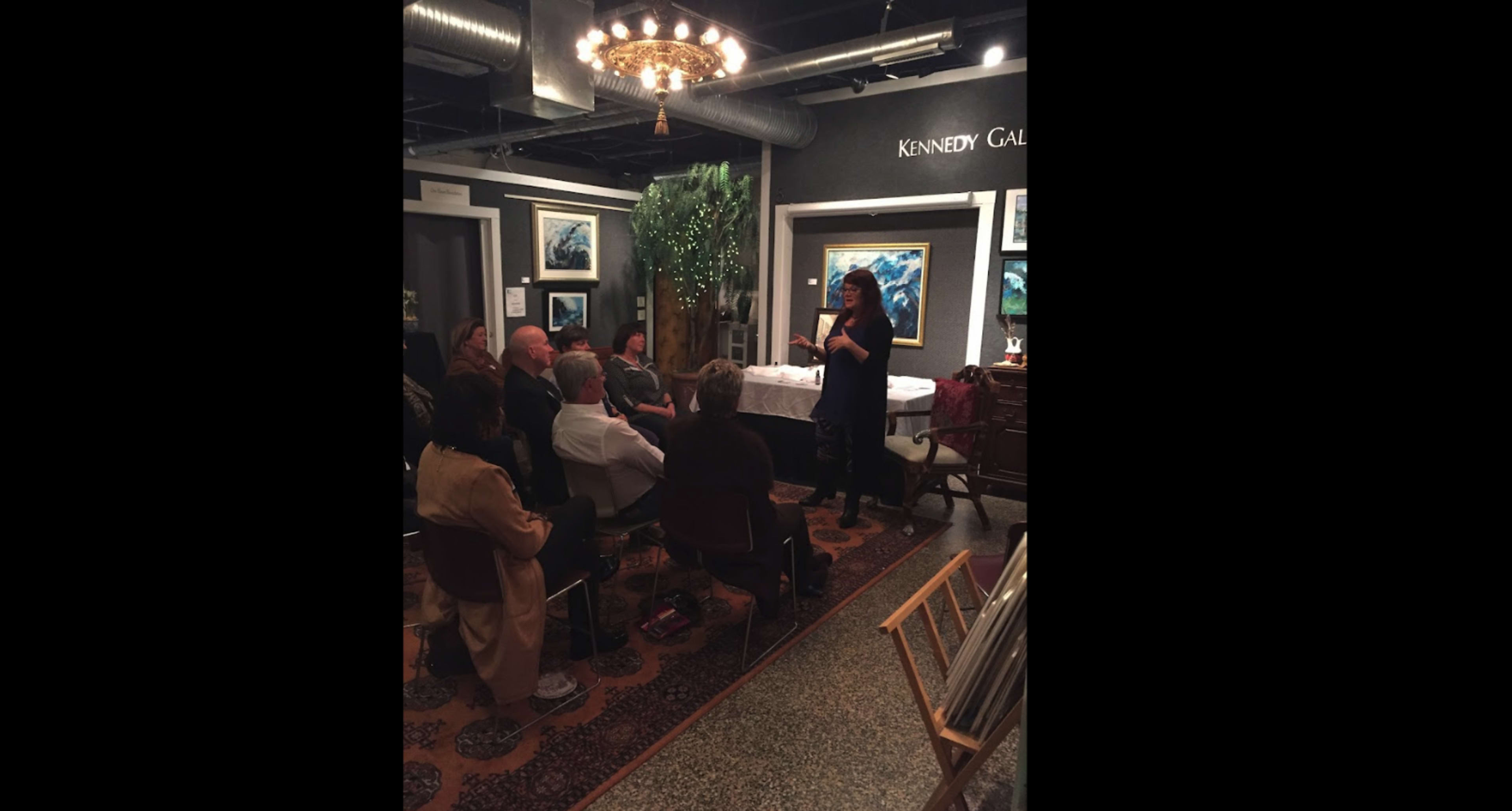 A speaker presents to an audience seated in a gallery adorned with artwork and plants.