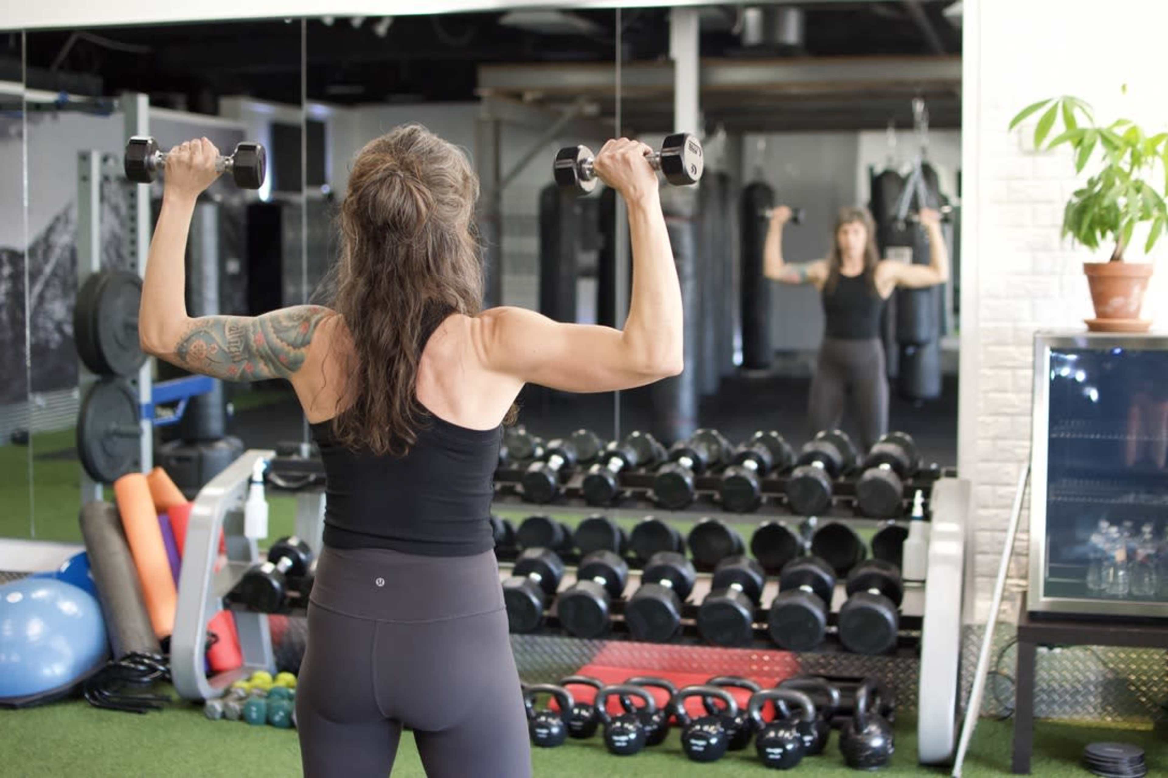 A person is lifting dumbbells in a gym while facing a mirror, with various fitness equipment visible in the background.