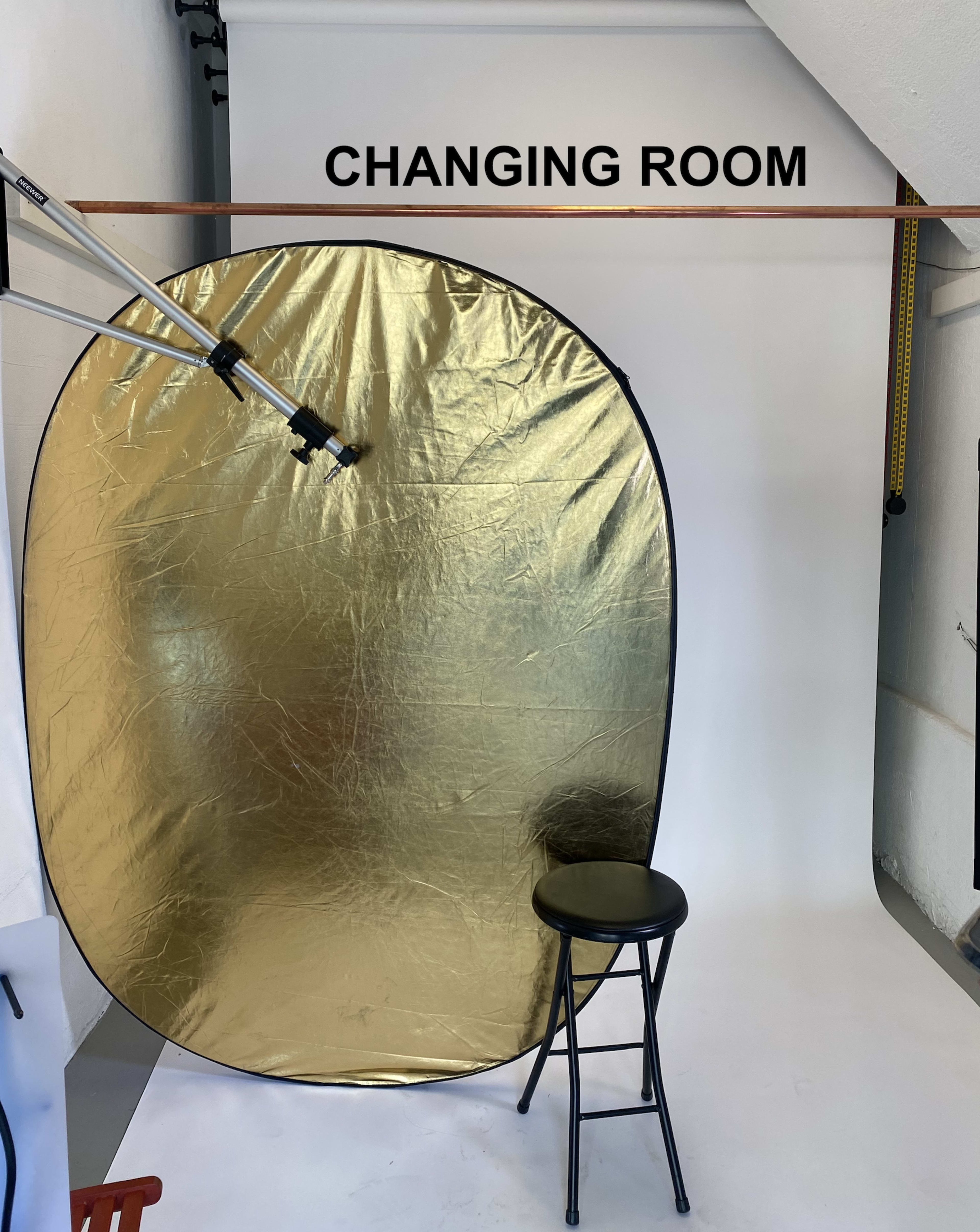 A changing room setup with a gold reflector, a stool, and a plain white backdrop.