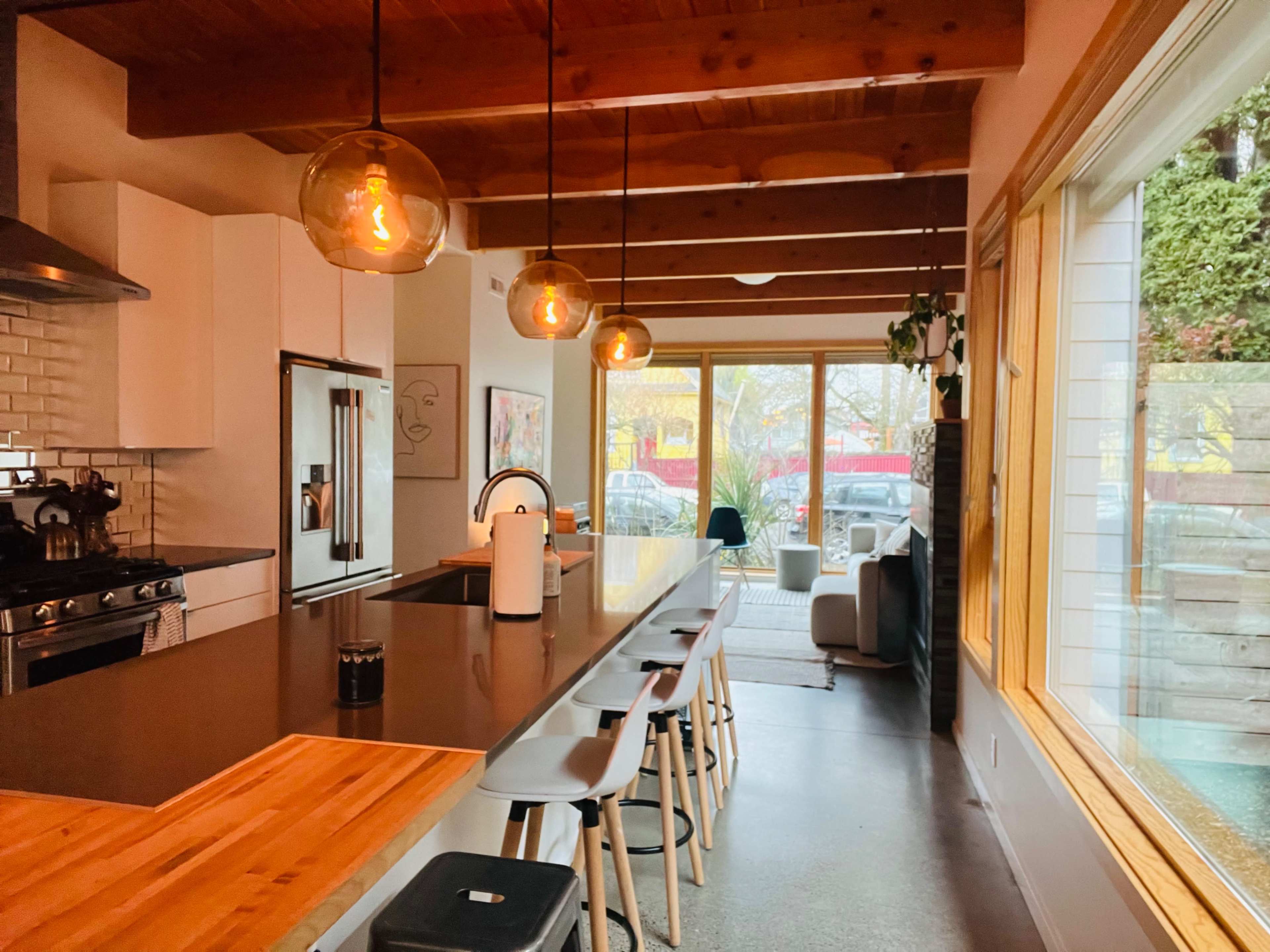 A modern kitchen features a large island with wooden and steel finishes, hanging pendant lights, and a spacious window providing natural light and a view of the outside.