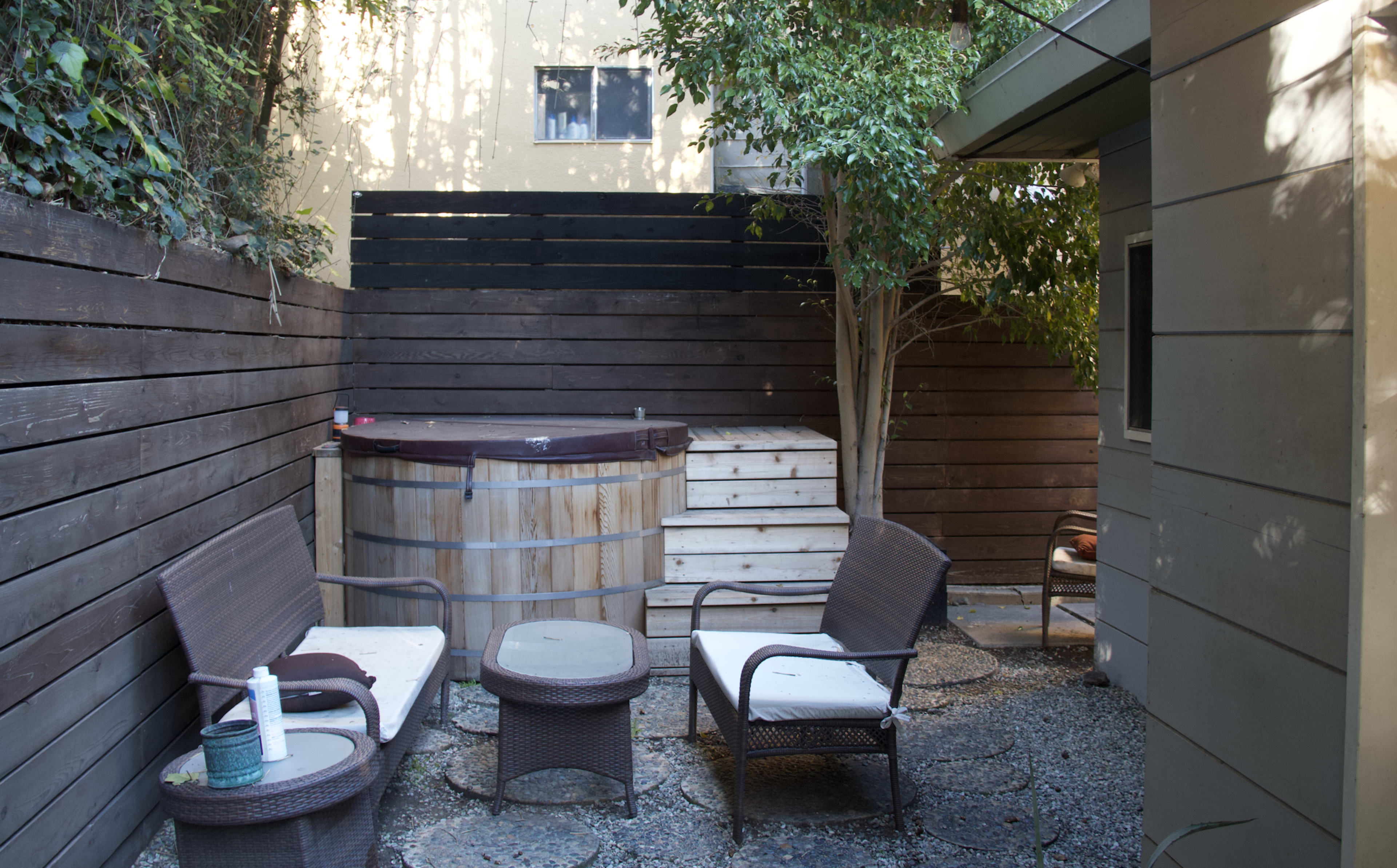 A small outdoor space featuring a wooden hot tub, two chairs, and a table surrounded by wooden fencing and greenery.