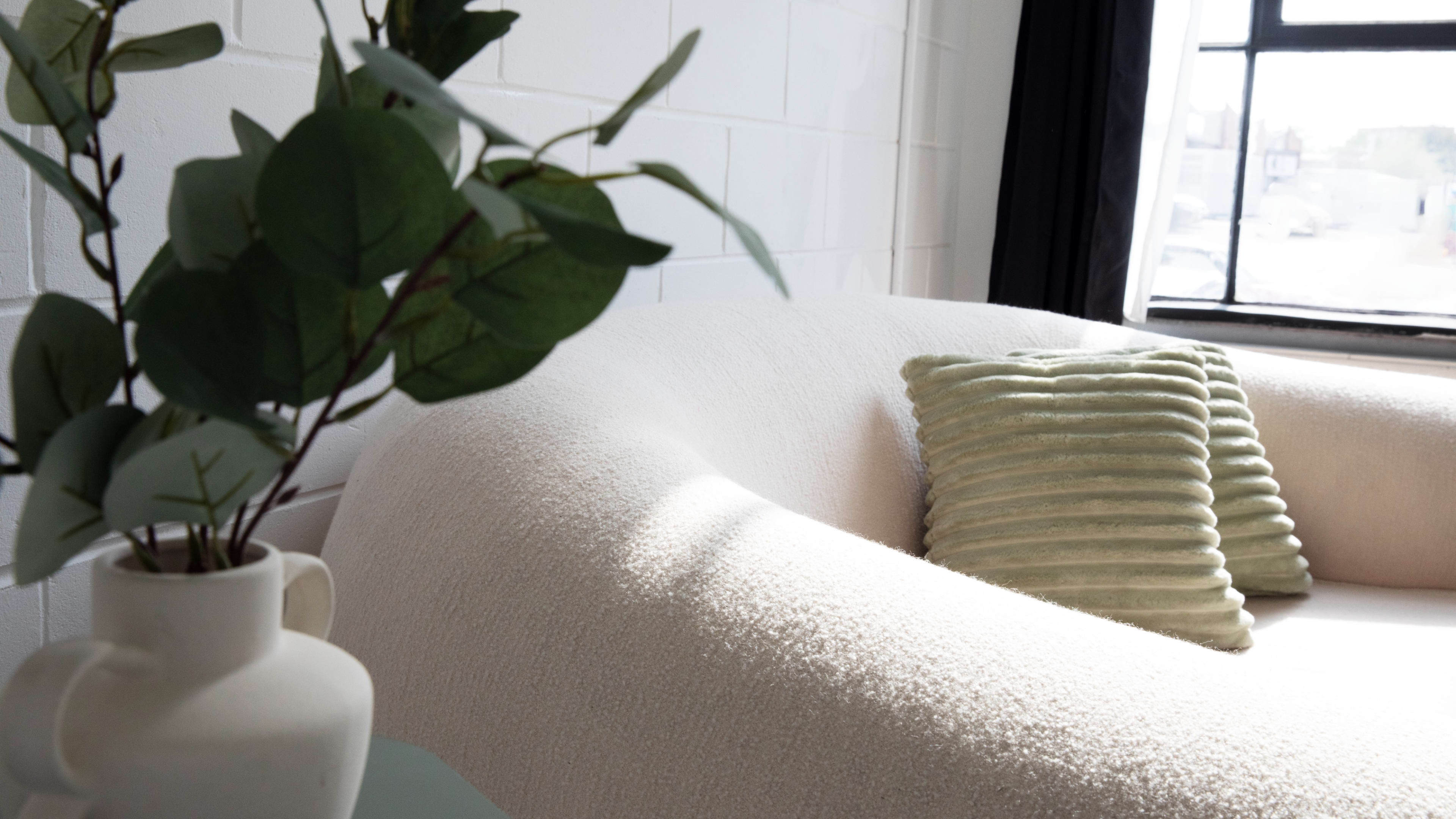 A plush, curved sofa is paired with a textured green pillow, beside a vase with leafy foliage, bathed in natural light from a nearby window.